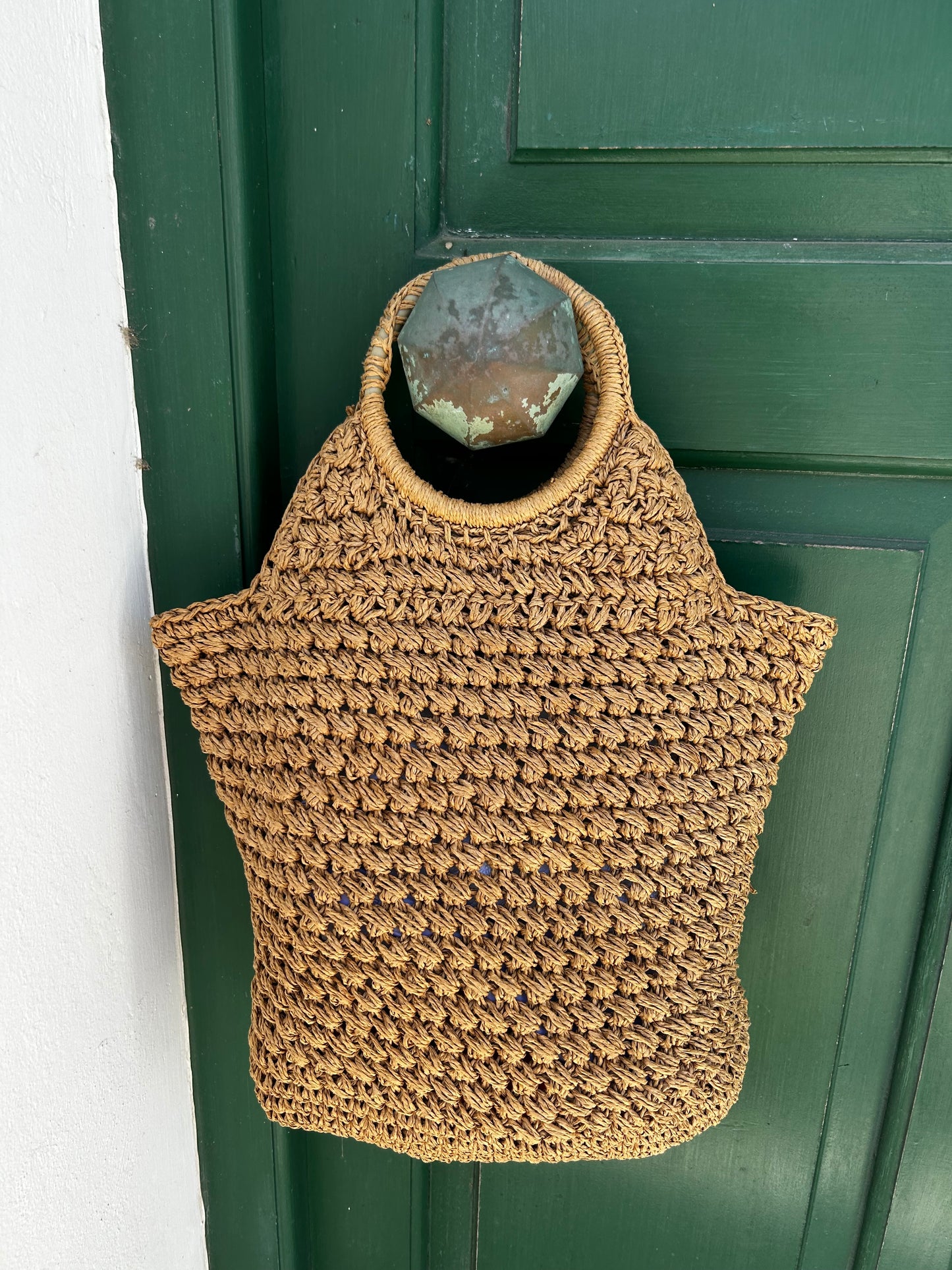 A hand-woven straw bag in a natural tan color, designed with a loop on the side to hang from a dark green wooden door.