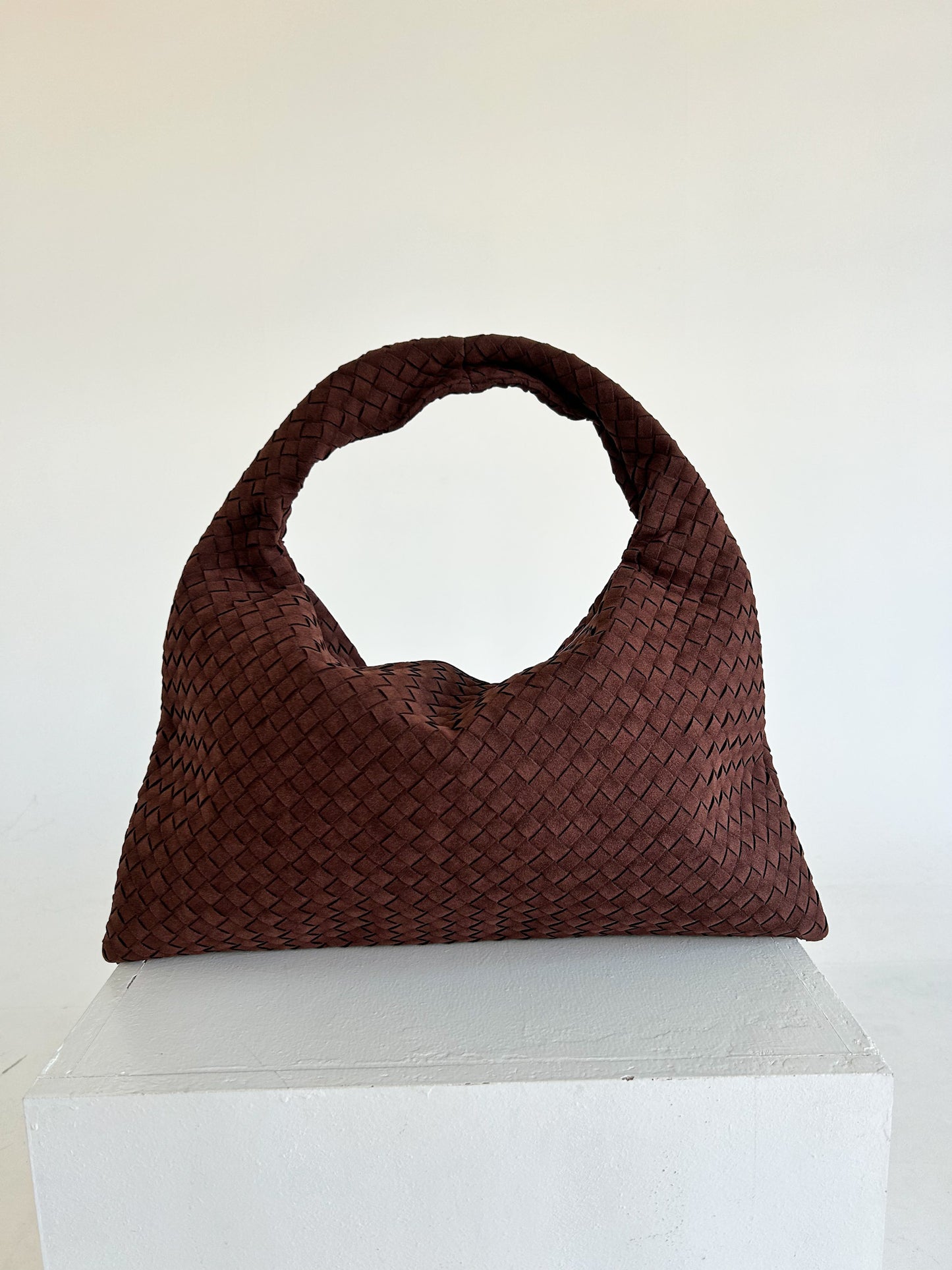 Brown woven handbag on a white pedestal against a white background