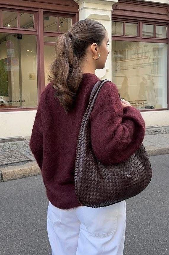 Woman in a burgundy sweater and white pants holding a textured brown handbag on a street.