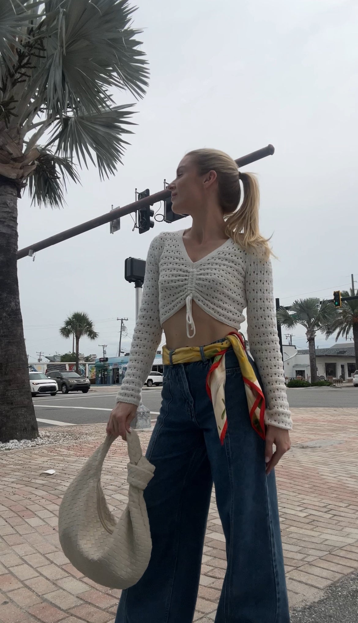 Woman in a white knitted cardigan and blue jeans holding a beige woven bag from PattyB on a street with palm trees.