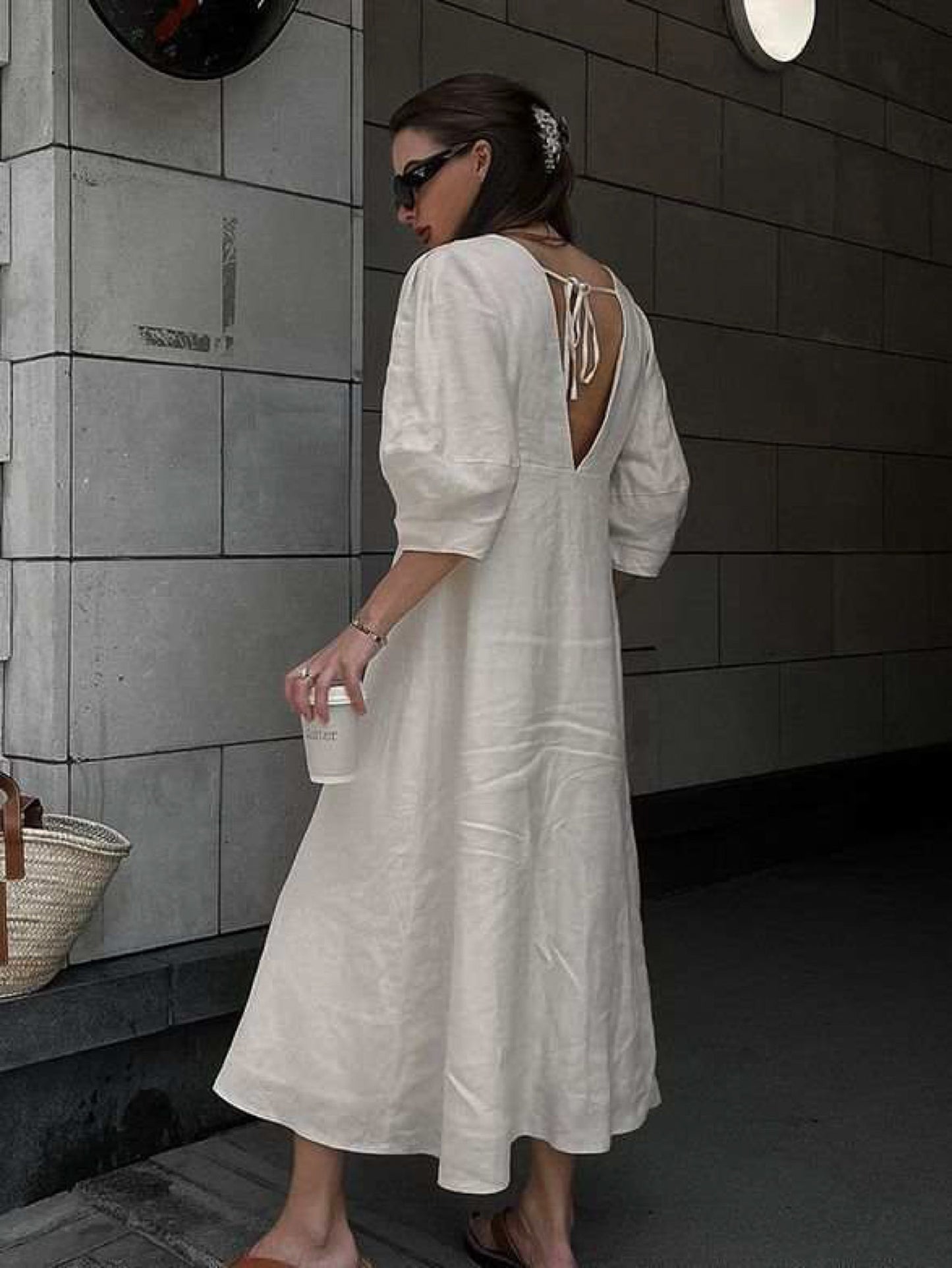 Back view of a woman wearing a cream linen midi dress featuring an open V-back with tie detail and elbow-length puff sleeves. The dress has a relaxed A-line silhouette and is styled with brown slide sandals and black sunglasses, creating a chic, effortless summer look.