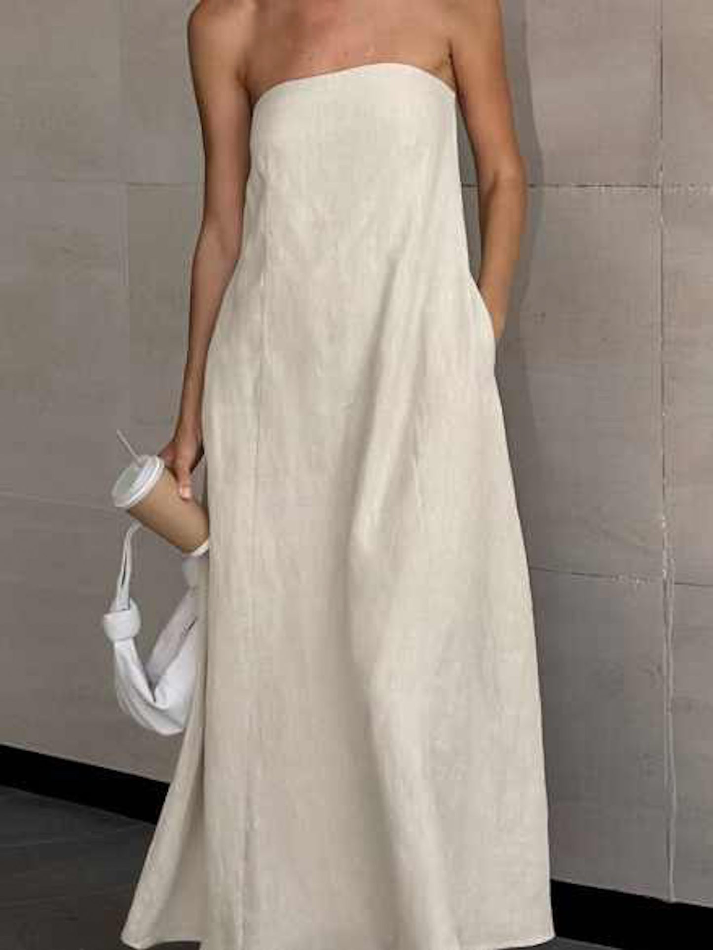 Woman wearing a minimalist strapless linen maxi dress in a neutral beige shade, styled with black sandals, statement sunglasses, and a white knotted handbag. The dress features a clean, structured bodice and flows into a relaxed, floor-length silhouette—perfect for warm-weather elegance and effortless style.