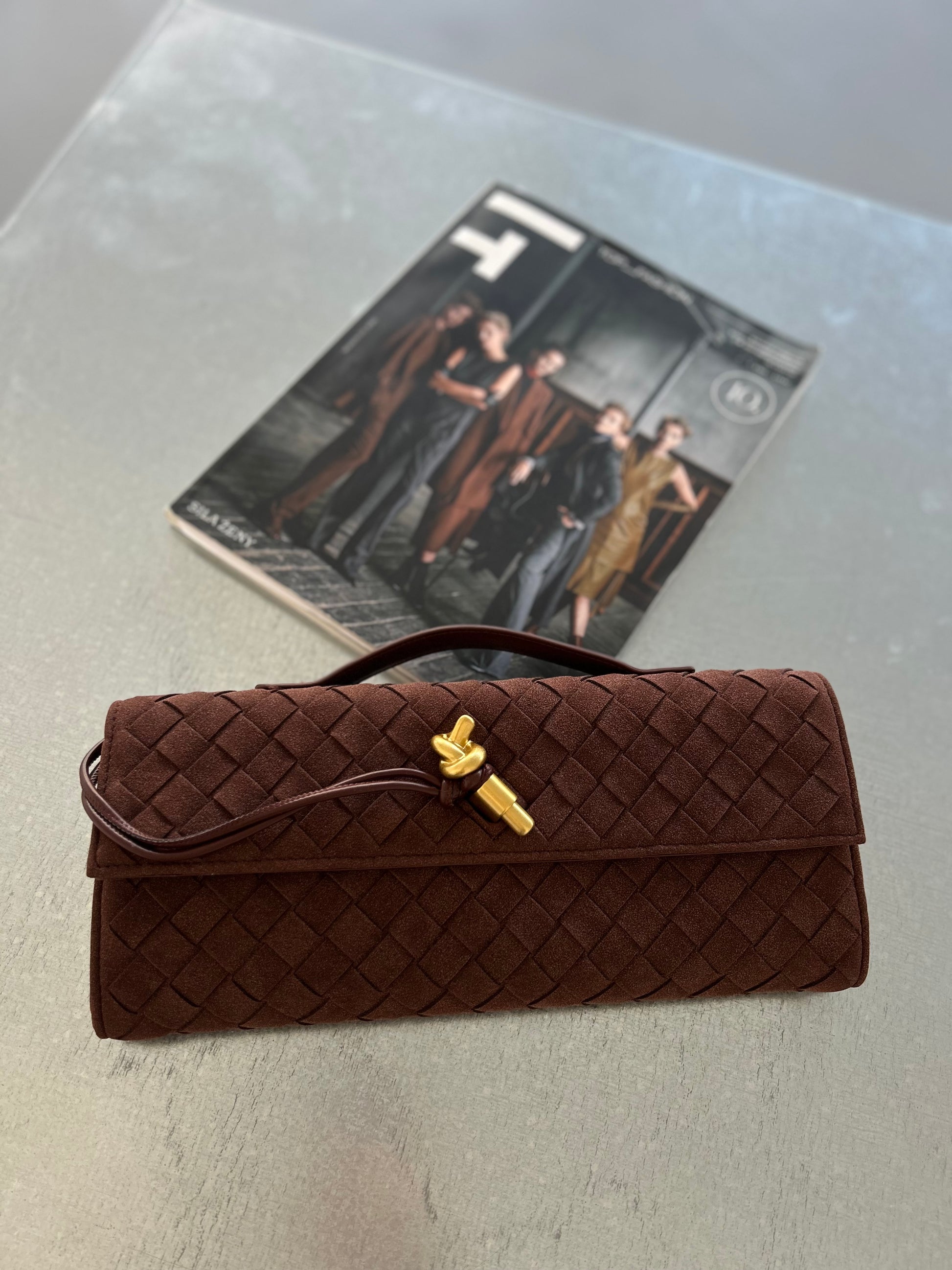 A brown suede card case handbag with a quilted design and a gold fastener, displayed on a surface with a magazine in the background.