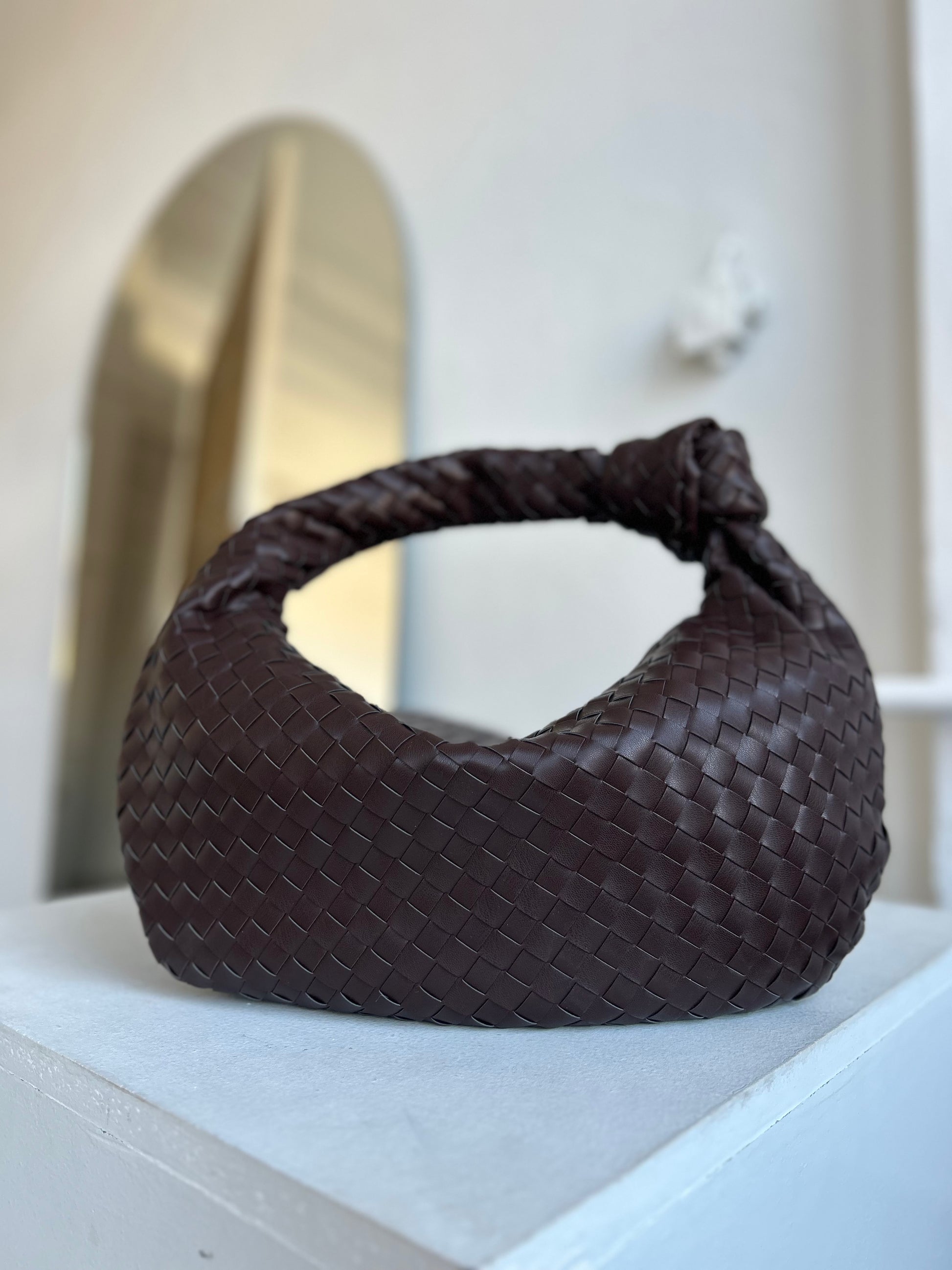 A brown woven dumpling-shaped shoulder handbag made of soft leather, displayed against an interior background.
