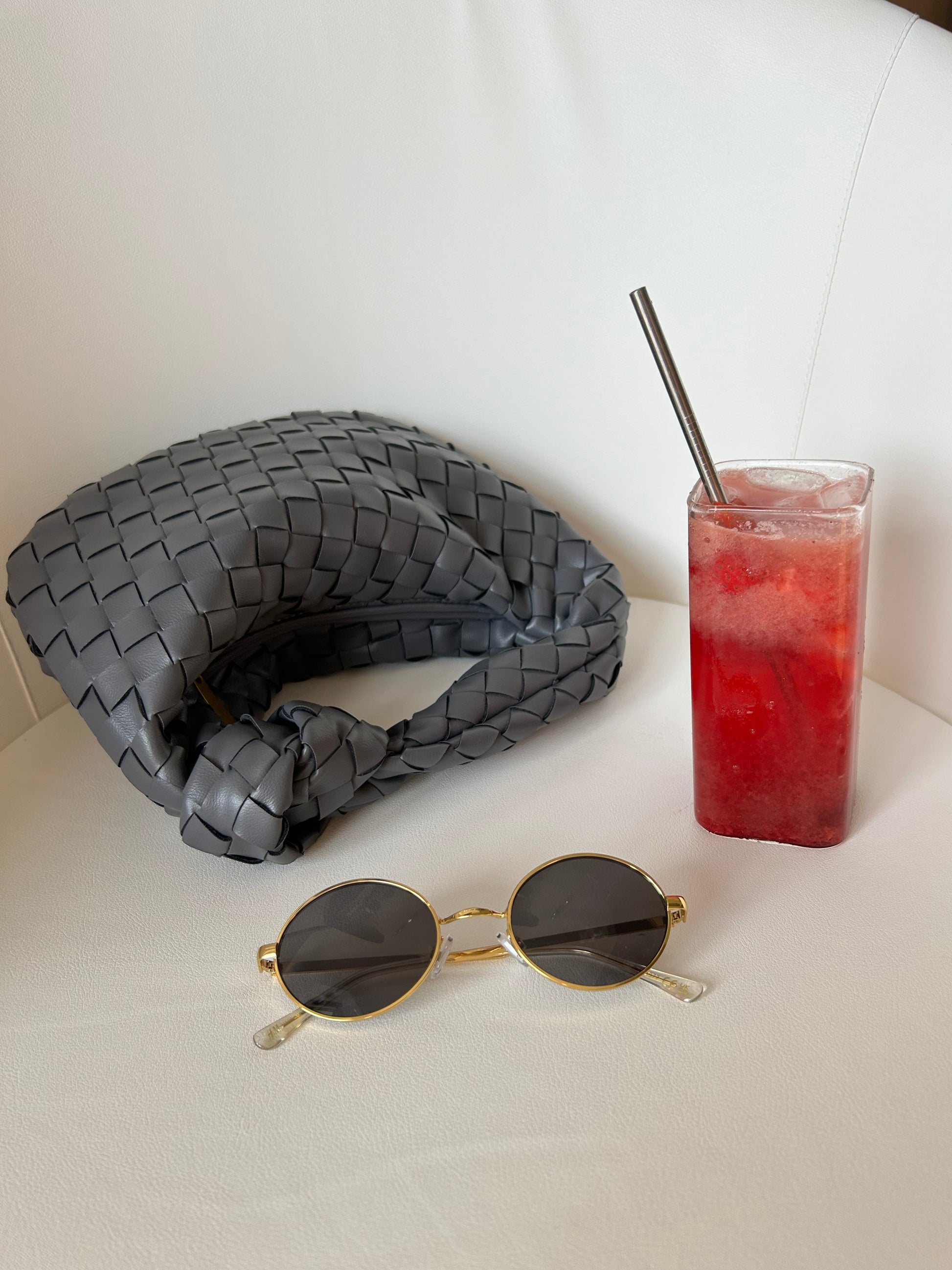 A gray woven clutch with a braided top handle, displayed next to a glass of red drink and a pair of sunglasses on a white surface.