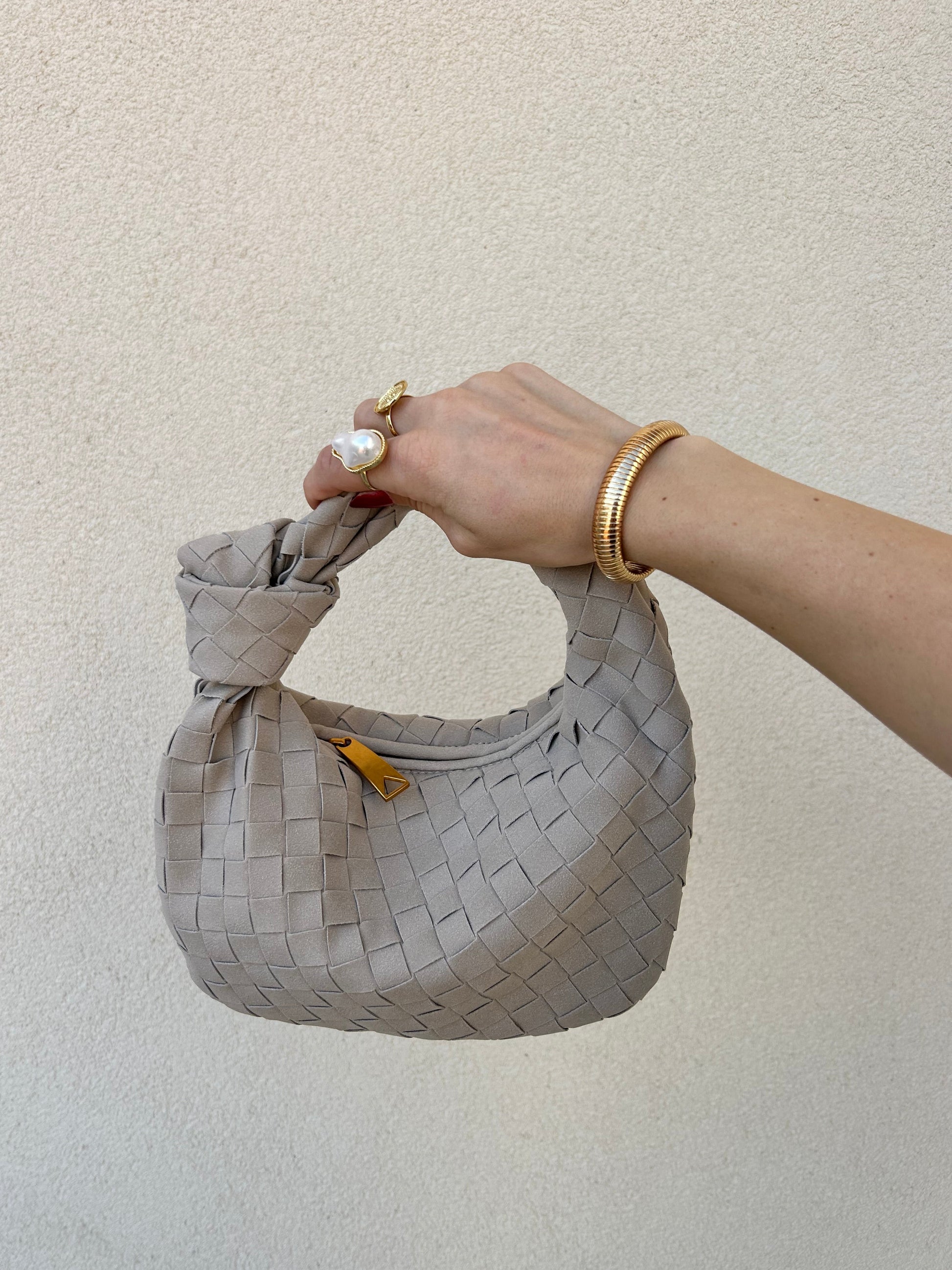 A hand holding a small, light grey vegan leather - suede clutch bag with a braided top handle and a gold hardware closure.