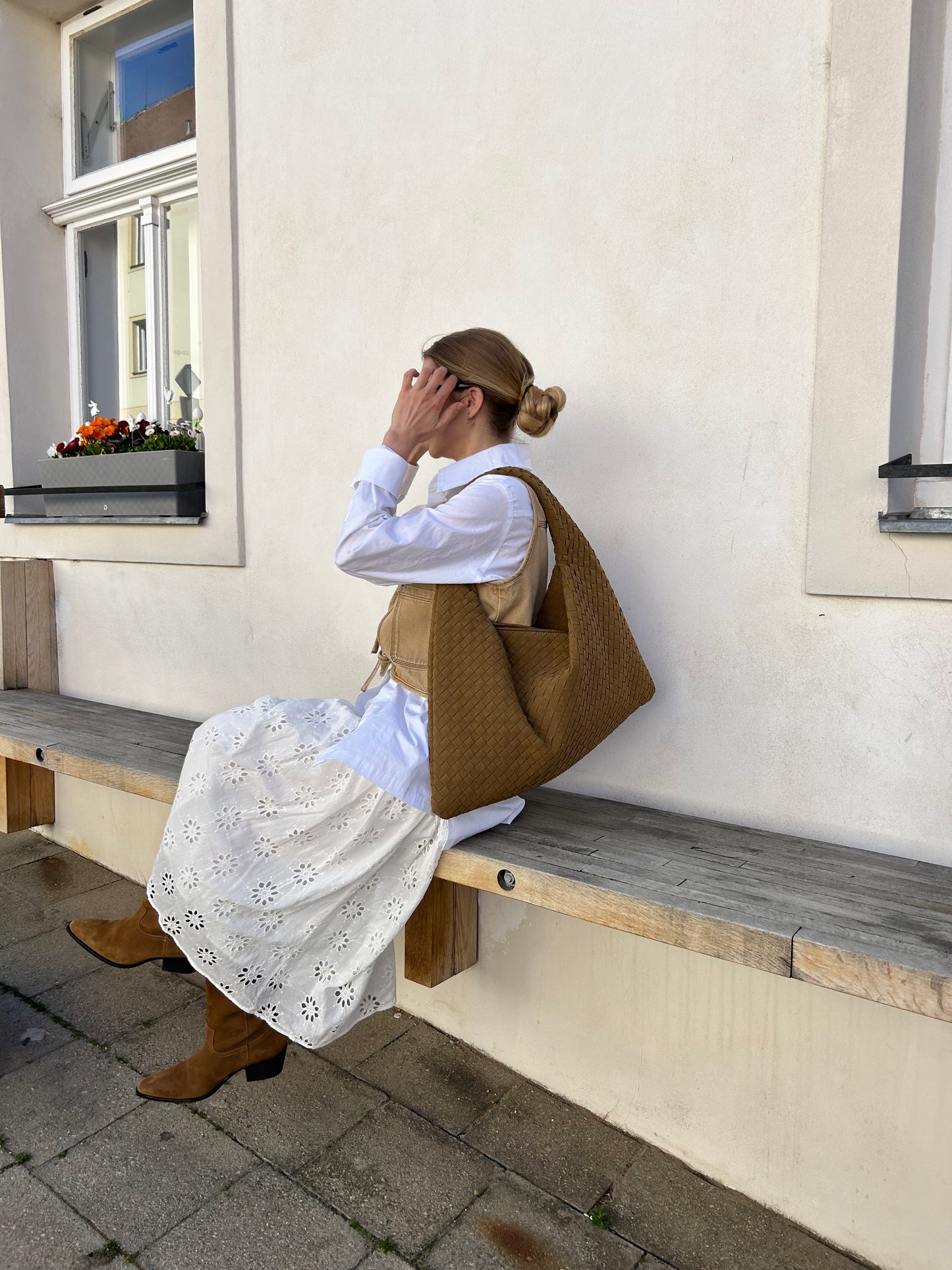 A woman seated on a bench wearing a white dress with a large woven design handbag on their shoulder.