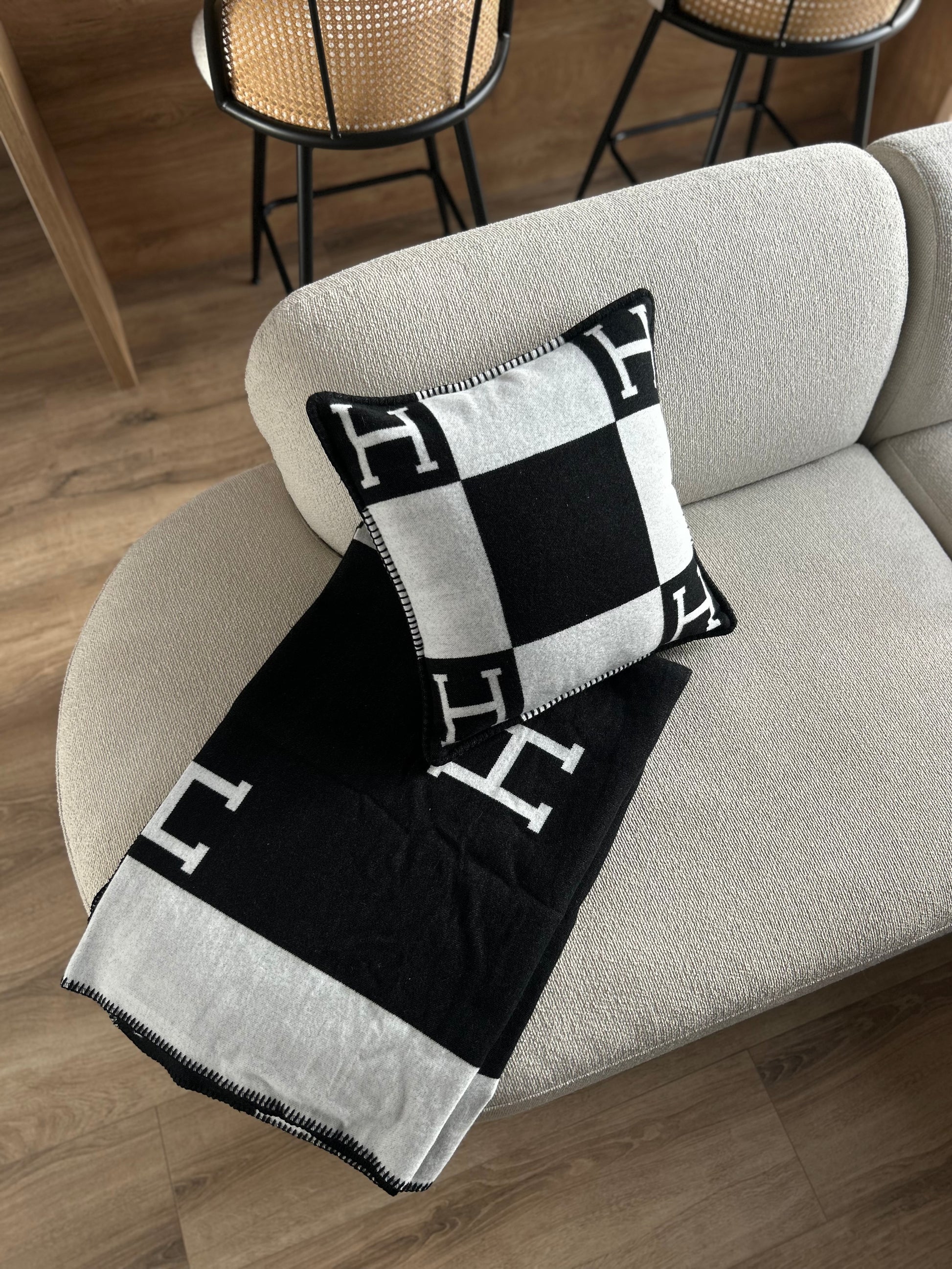 Black and white patterned pillow and throw blanket on a beige sofa with wooden floor and chairs in the background.