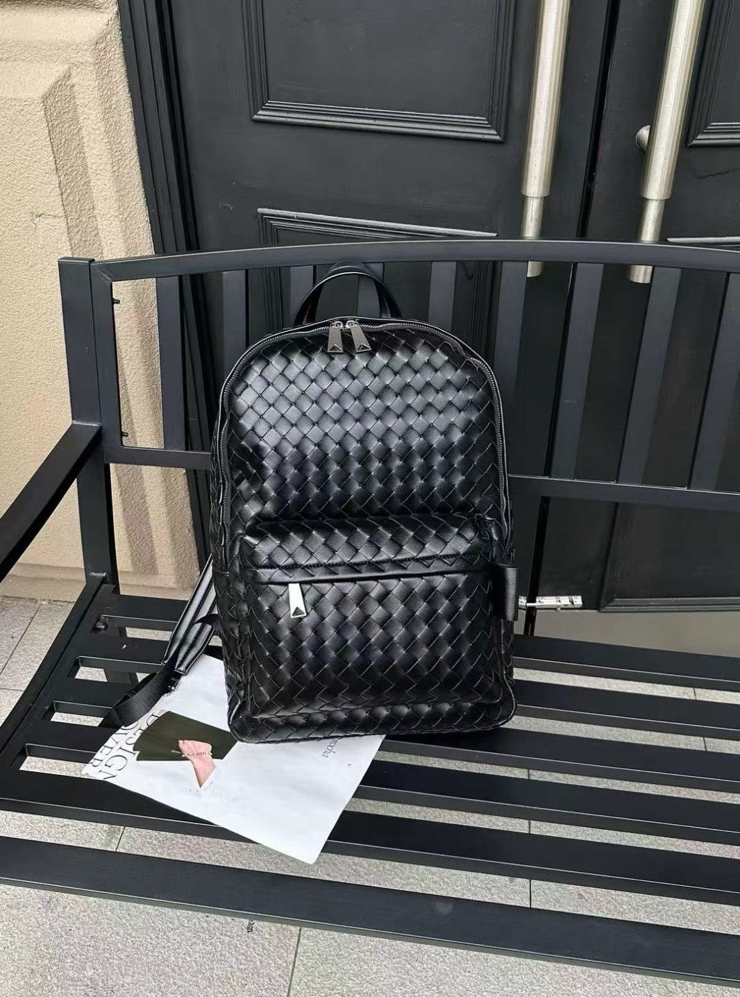 Black woven textured backpack on a black metal bench with a door in the background. Black woven cowhide leather unisex backpack with minimalist luxury design perfect for work, travel and everyday use.