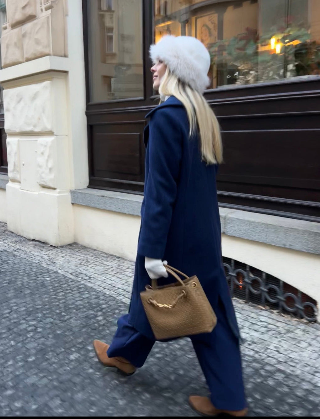 Blonde Slavic woman in a blue coat and white fur hat walking on a street of central Prague with a historical building in the background