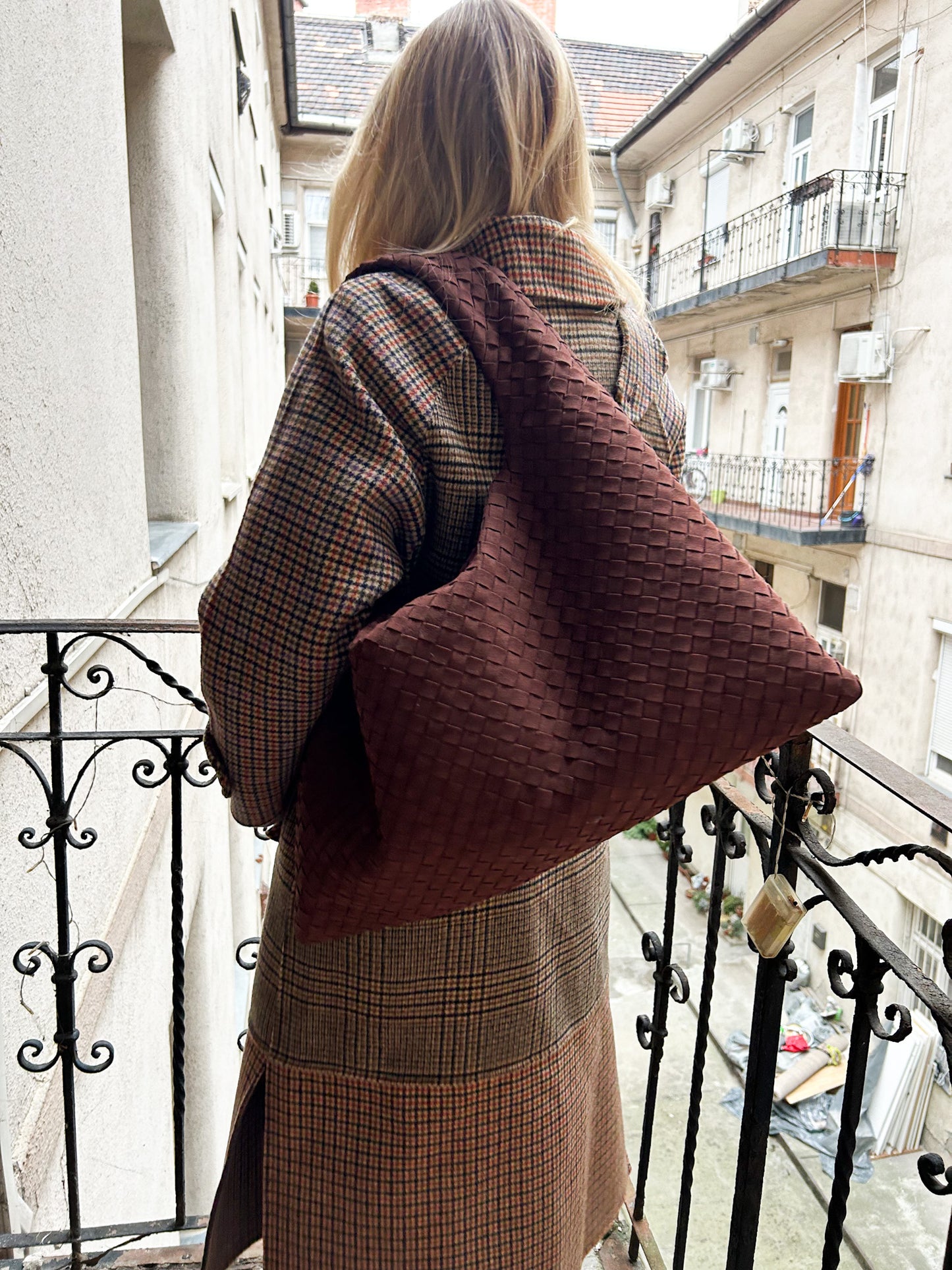 Blonde woman wearing a large brown woven handbag on a balcony with buildings in the background