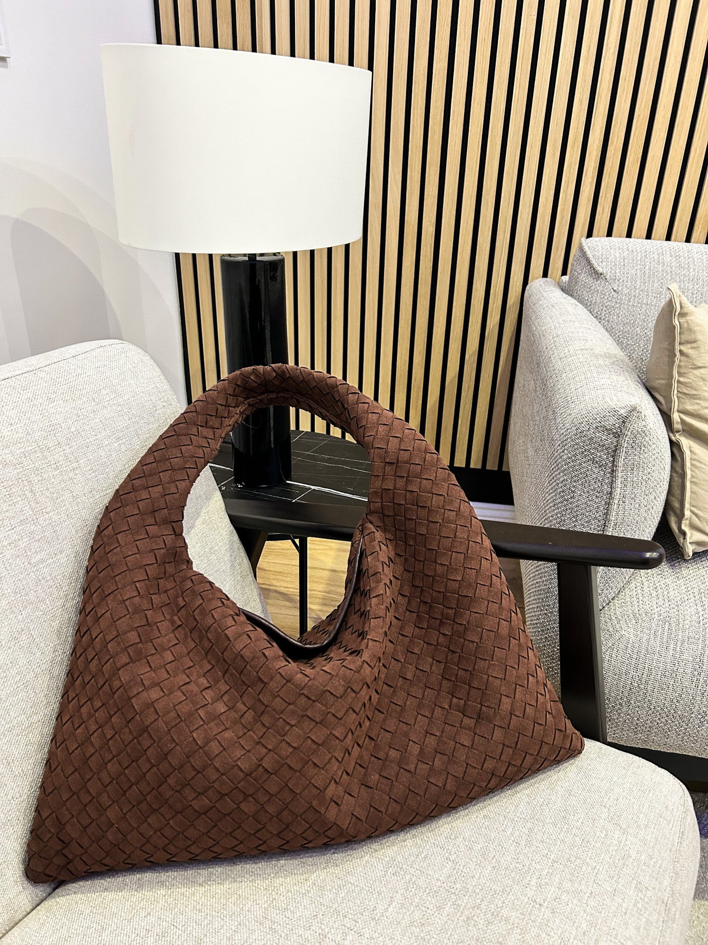 Brown woven handbag on a white sofa with a lamp and decorative pillows in the background.