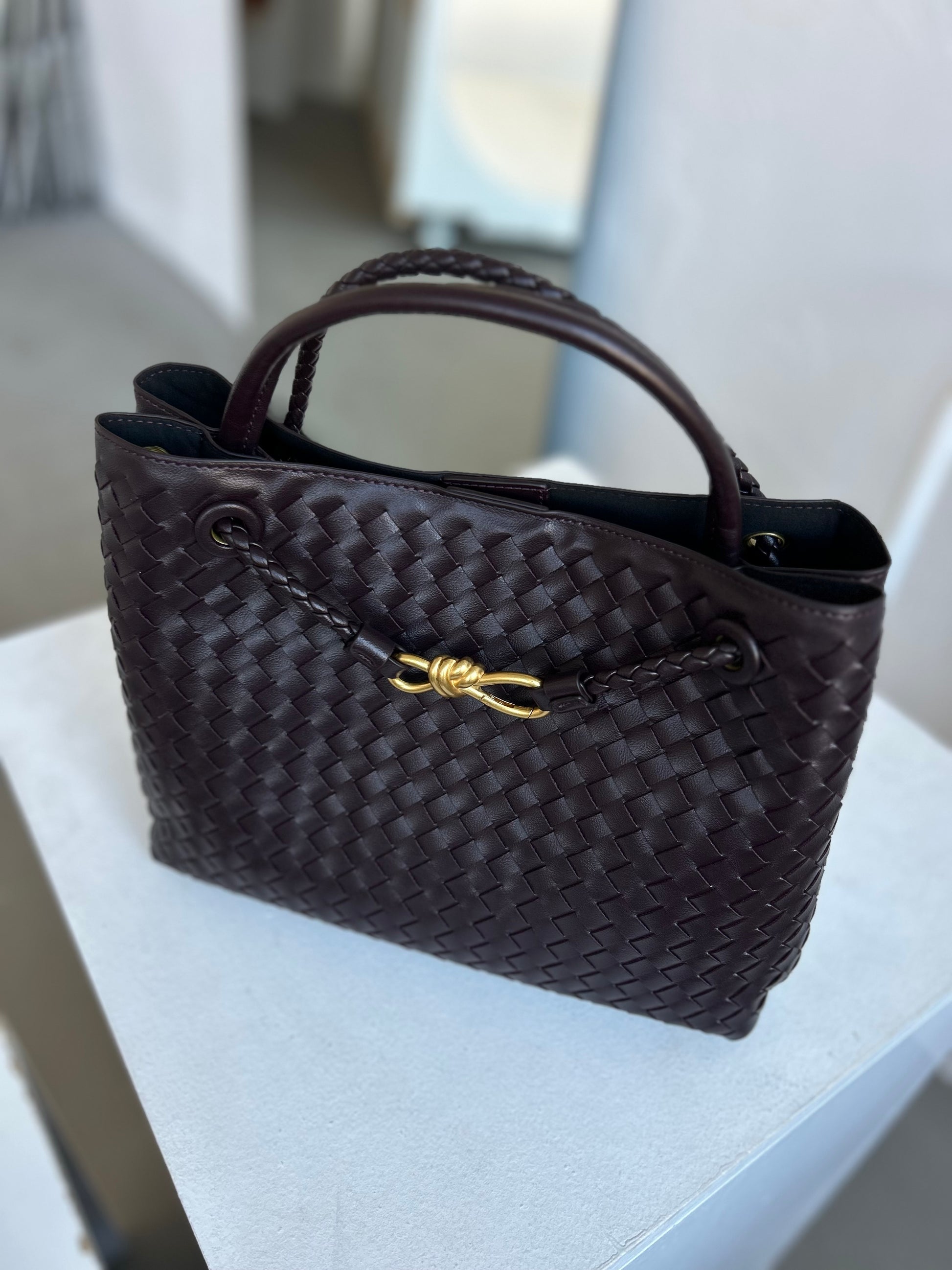 Dark chocolate woven vegan leather handbag with gold twist detail placed on a white chair