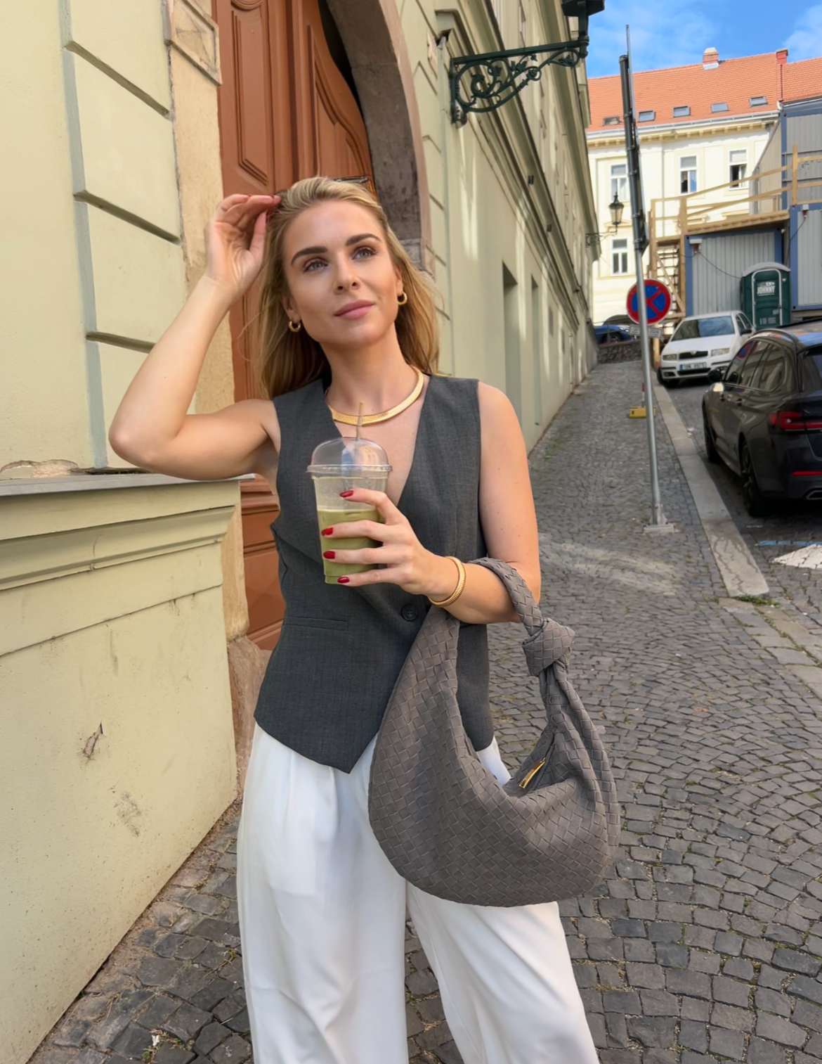 Blonde woman wearing summer outfit consisting of white wide leg pants and grey waistcoat, holding a grey woven knot bag with gold zipper. Interwoven shoulder hobo bag for everyday carry 