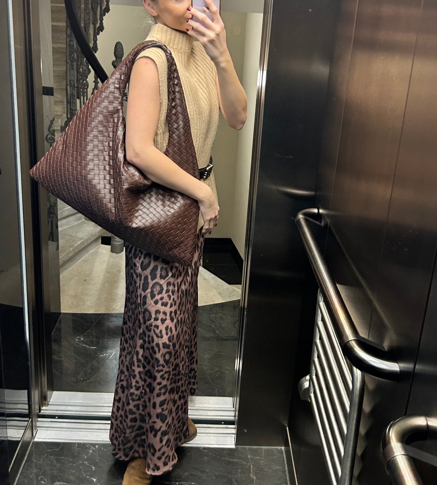 A stylish woman stands in  modern elevator,holding a stylish brown 
woven slouchy tote over her shoulder.The elegant accessory complements her outfit consisting of beige knitted tank top, black waist belt, leopard print skirt and brown boots to match