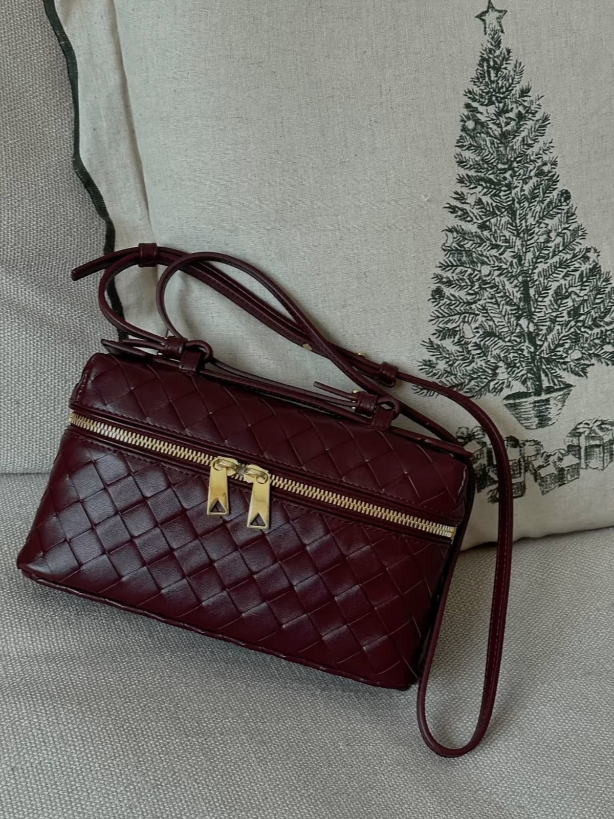Maroon woven handbag with gold zipper on a cream sofa, on a christmas pillow.Structured burgundy red crossbody bag with gold hardware