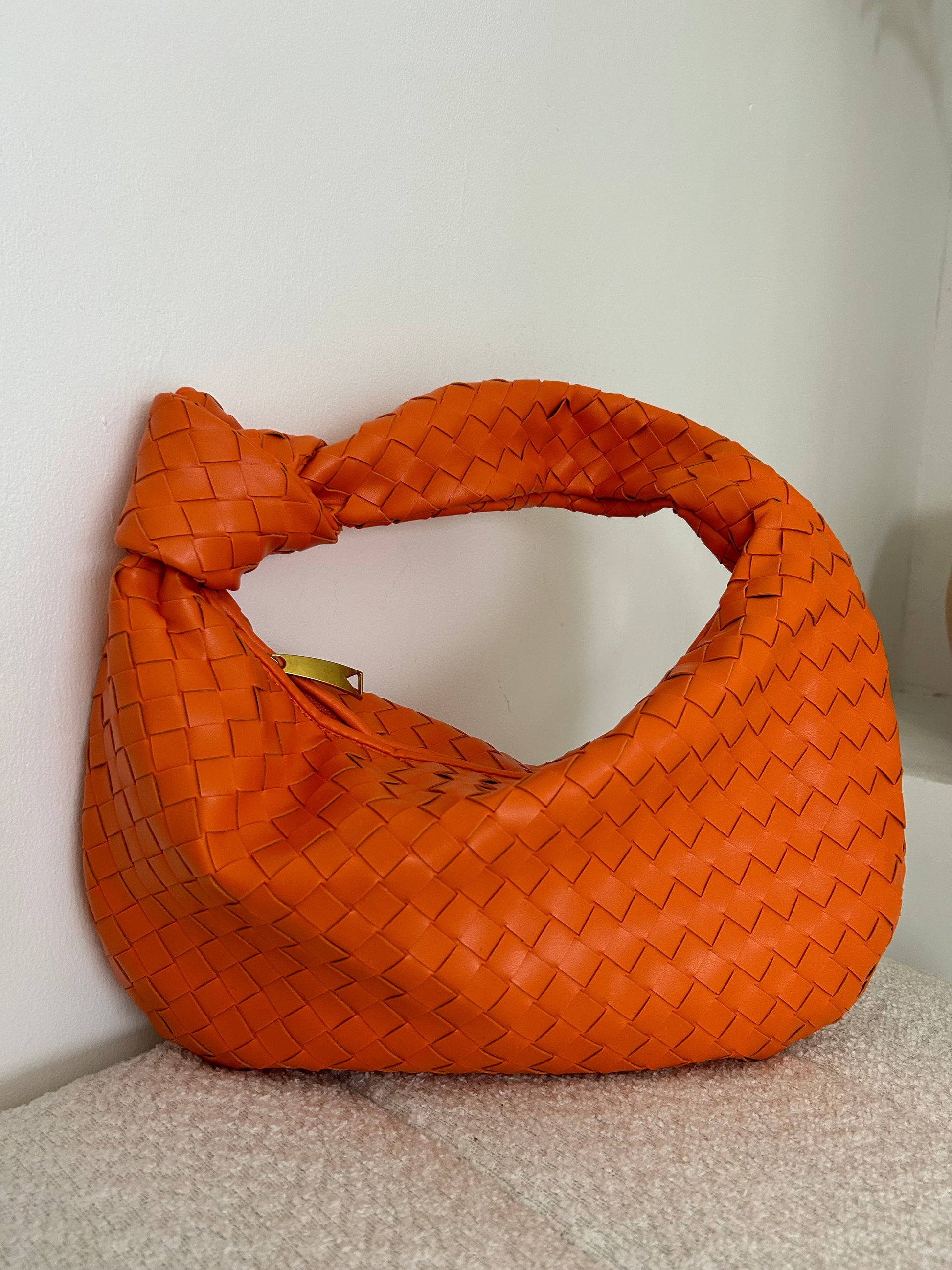 Close-up of a bright orange leather shoulder bag featuring a woven design and unique triangle zipper closure, styled on a decorative stairway. Perfect mix of craftsmanship and trend.