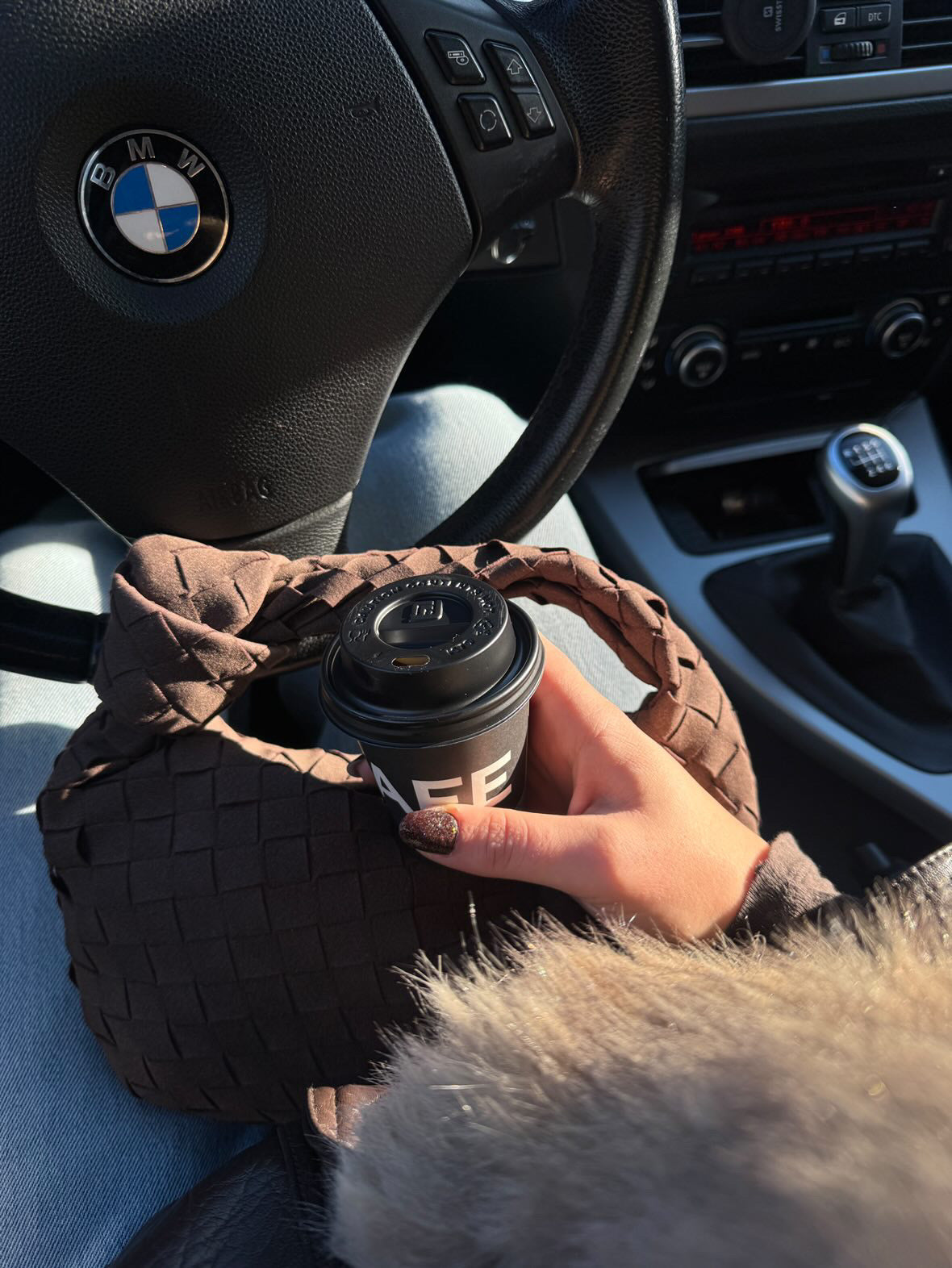 Person holding a coffee cup with a BMW steering wheel in the background. Soft vegan suede teddy clutch bag in chocolate brown