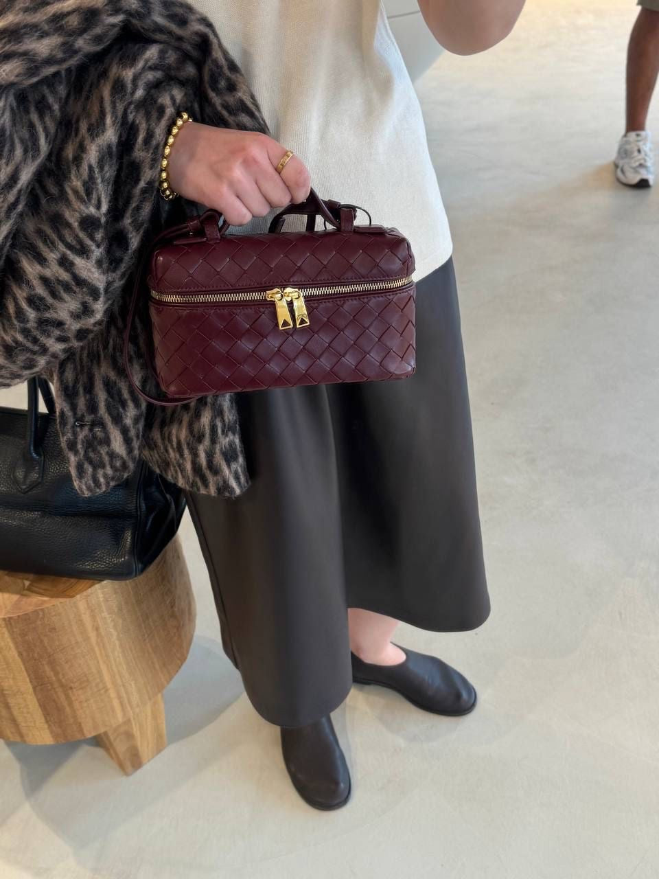 Person holding a maroon handbag with gold accents indoors. wearing an animal print coat, black trousers and black loafers