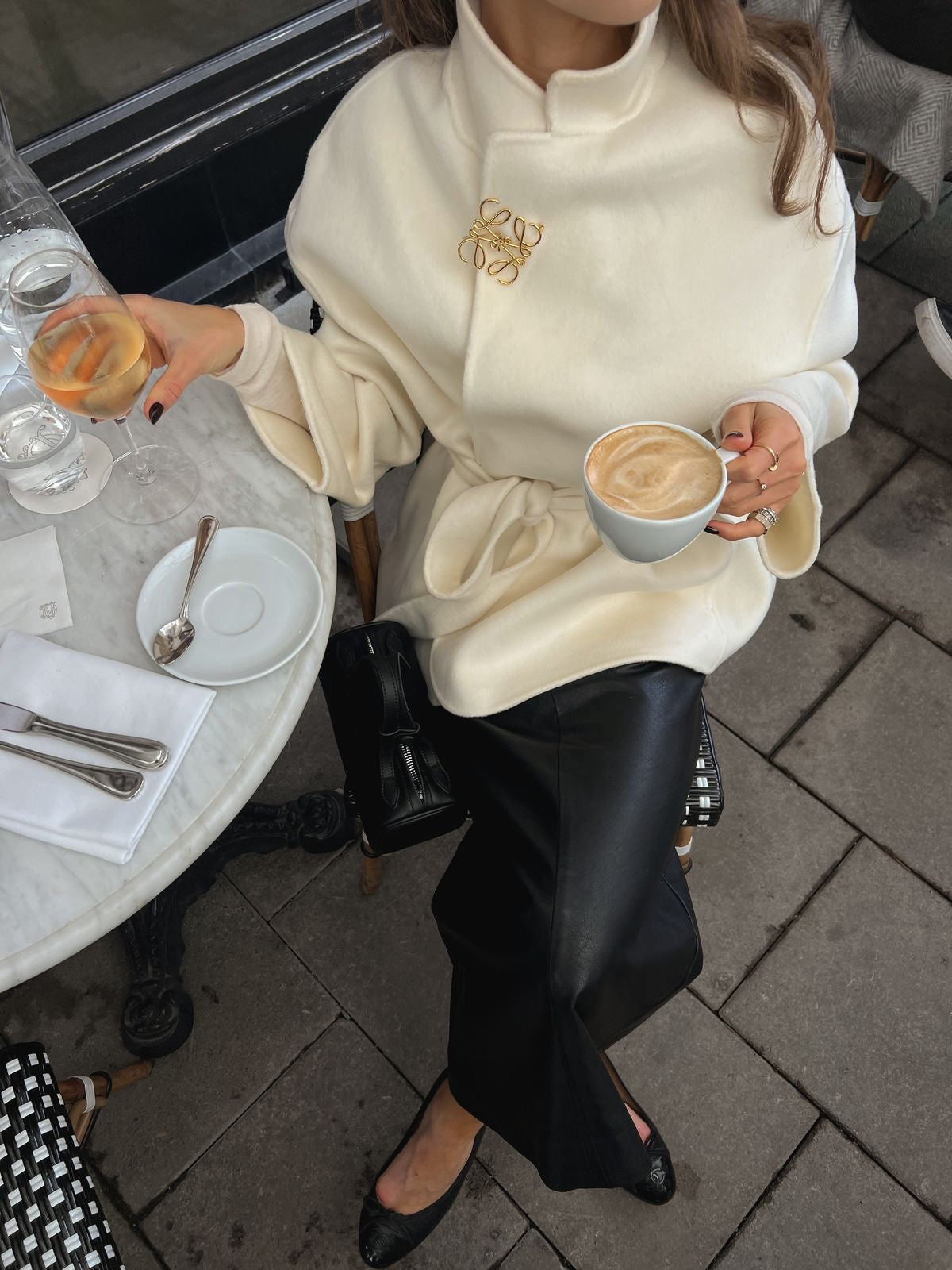 Person in a white coat with a gold brooch, holding a coffee cup outdoors.