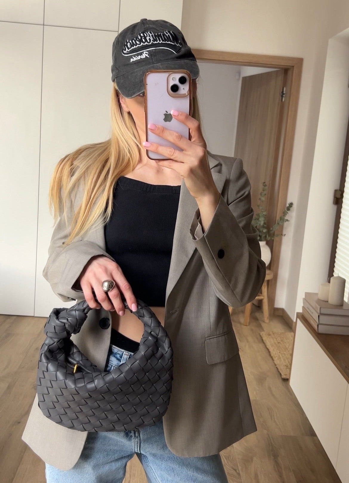 Person taking a mirror selfie wearing a cap, holding a phone, and carrying a textured bag indoors.