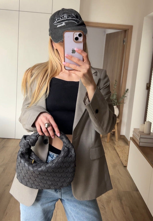 Person taking a mirror selfie wearing a cap, holding a phone, and carrying a textured bag indoors.