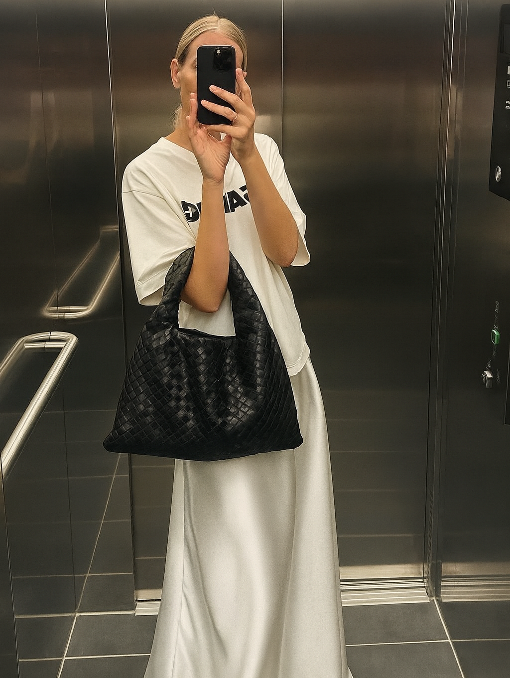 Person taking a mirror selfie wearing a white t-shirt and long white skirt, holding a black woven bag.