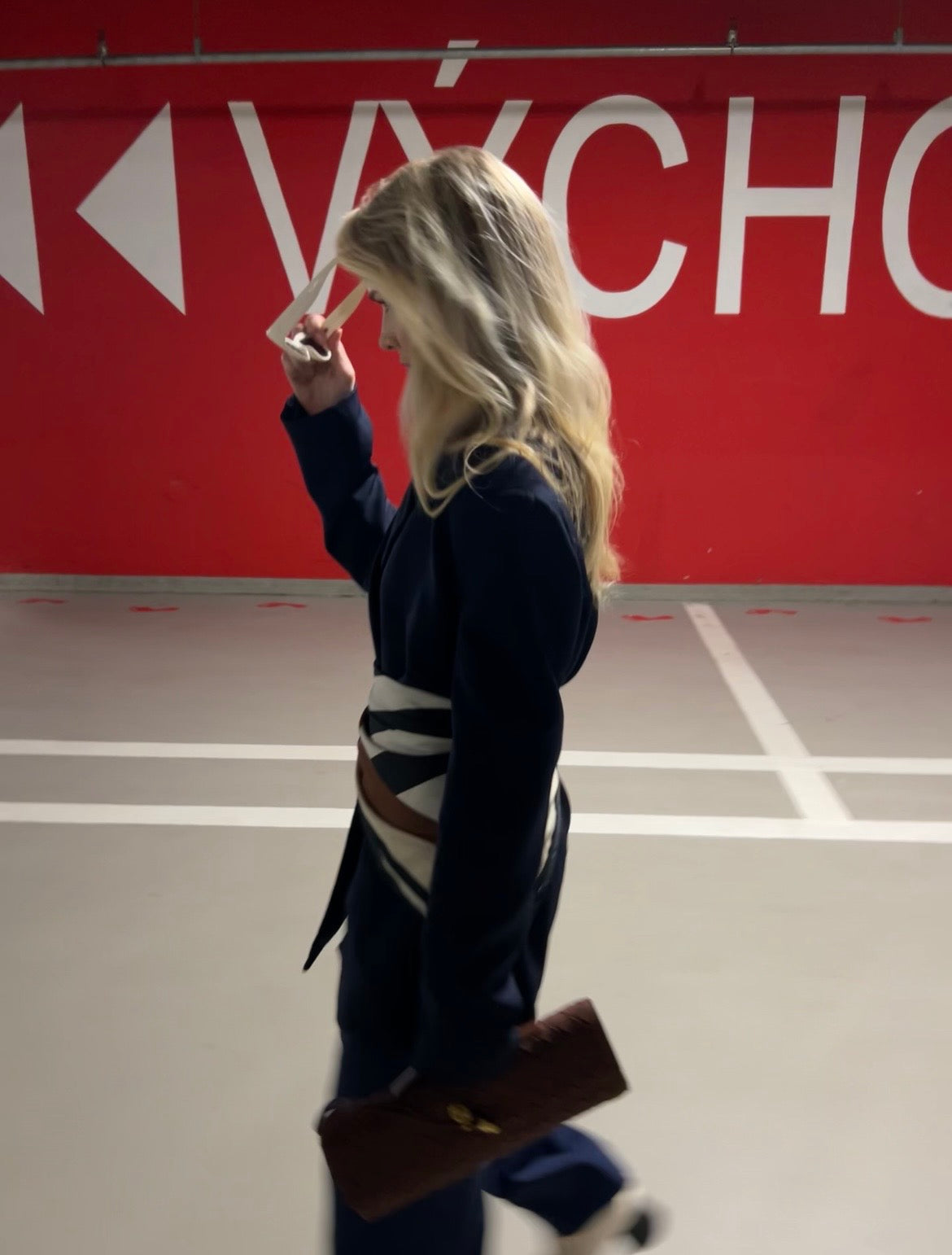 Person walking in a parking lot with a red wall and white text in the background, wearing a navy blue outfit, sunglasses and brown vegan suede clutch.