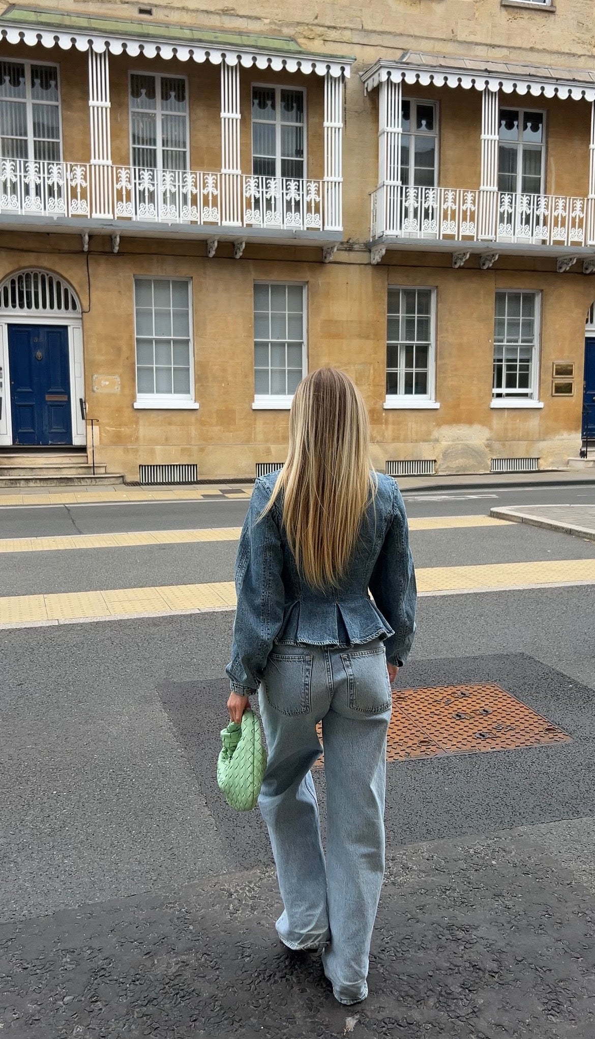Person walking on a street in Oxford in front of a building with a blue door wearing denim outfit with sage green woven knot bag.