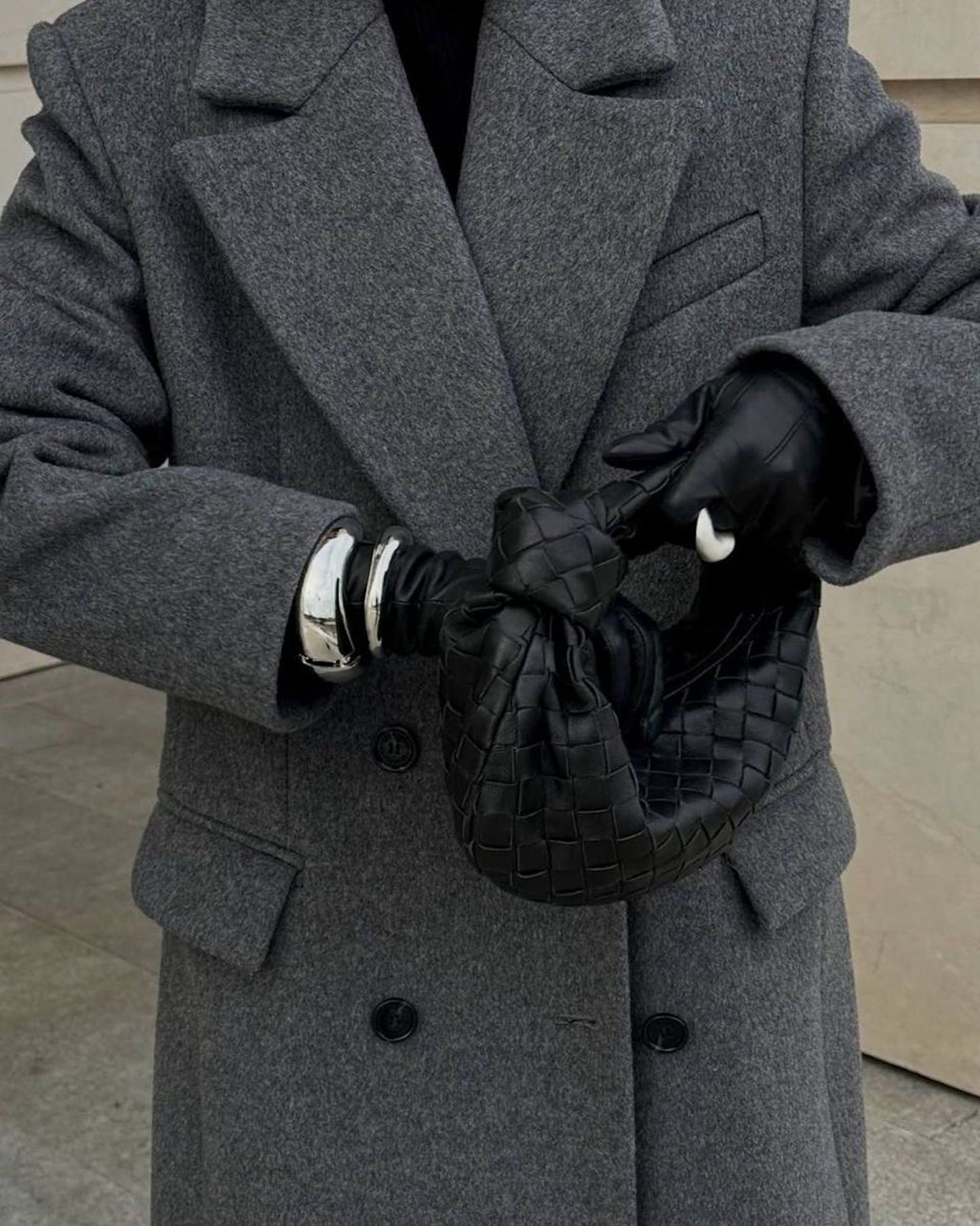 Person wearing a grey coat with black gloves and a woven designer inspired black bag.