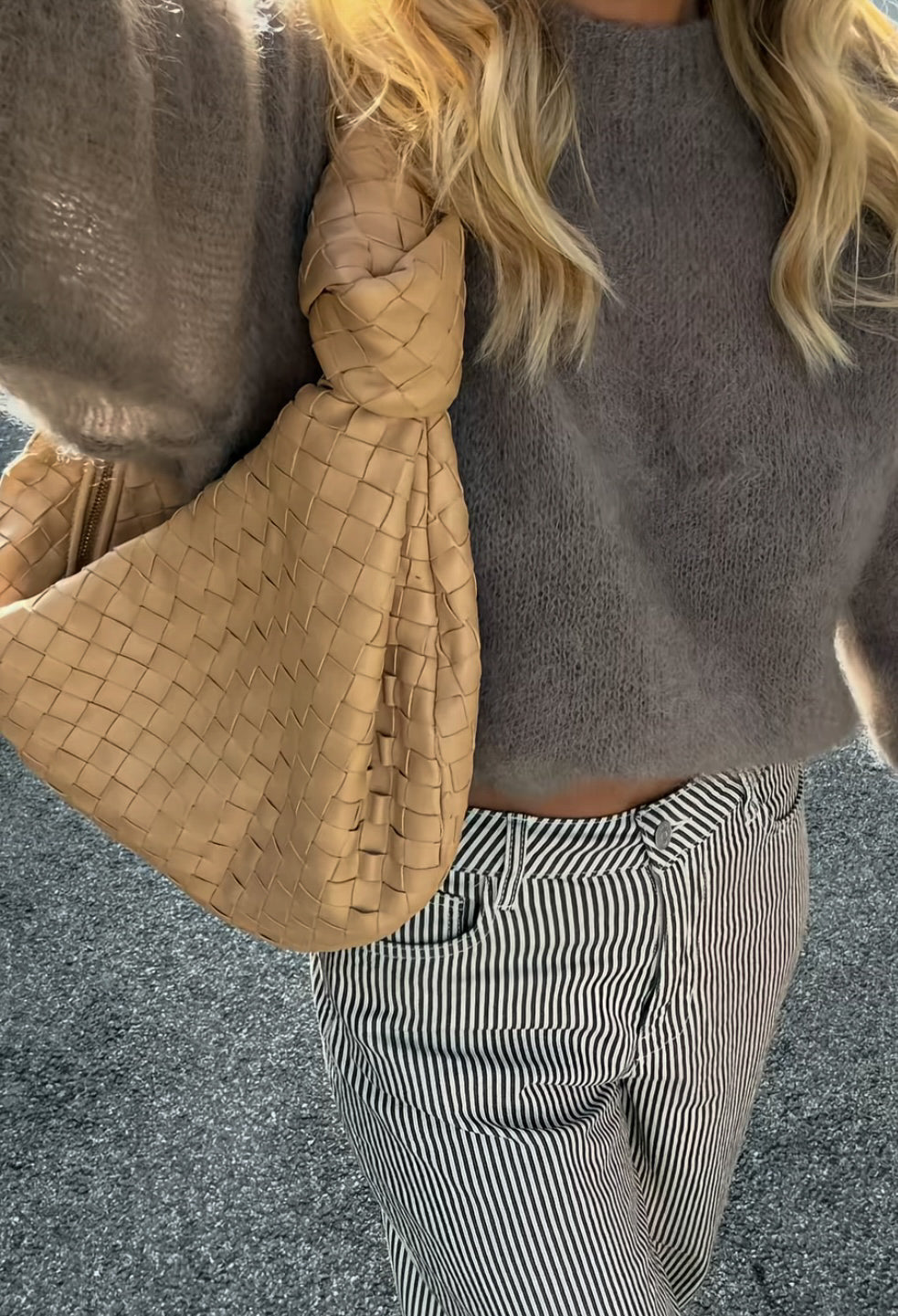 Person wearing a grey sweater and striped pants, holding a woven beige bag on her shoulder