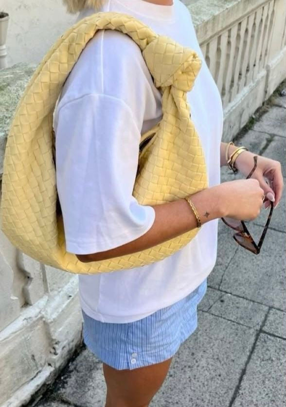 Person wearing a yellow woven designer inspired bag, white shirt, blue shorts, and yellow shoes on a sidewalk.