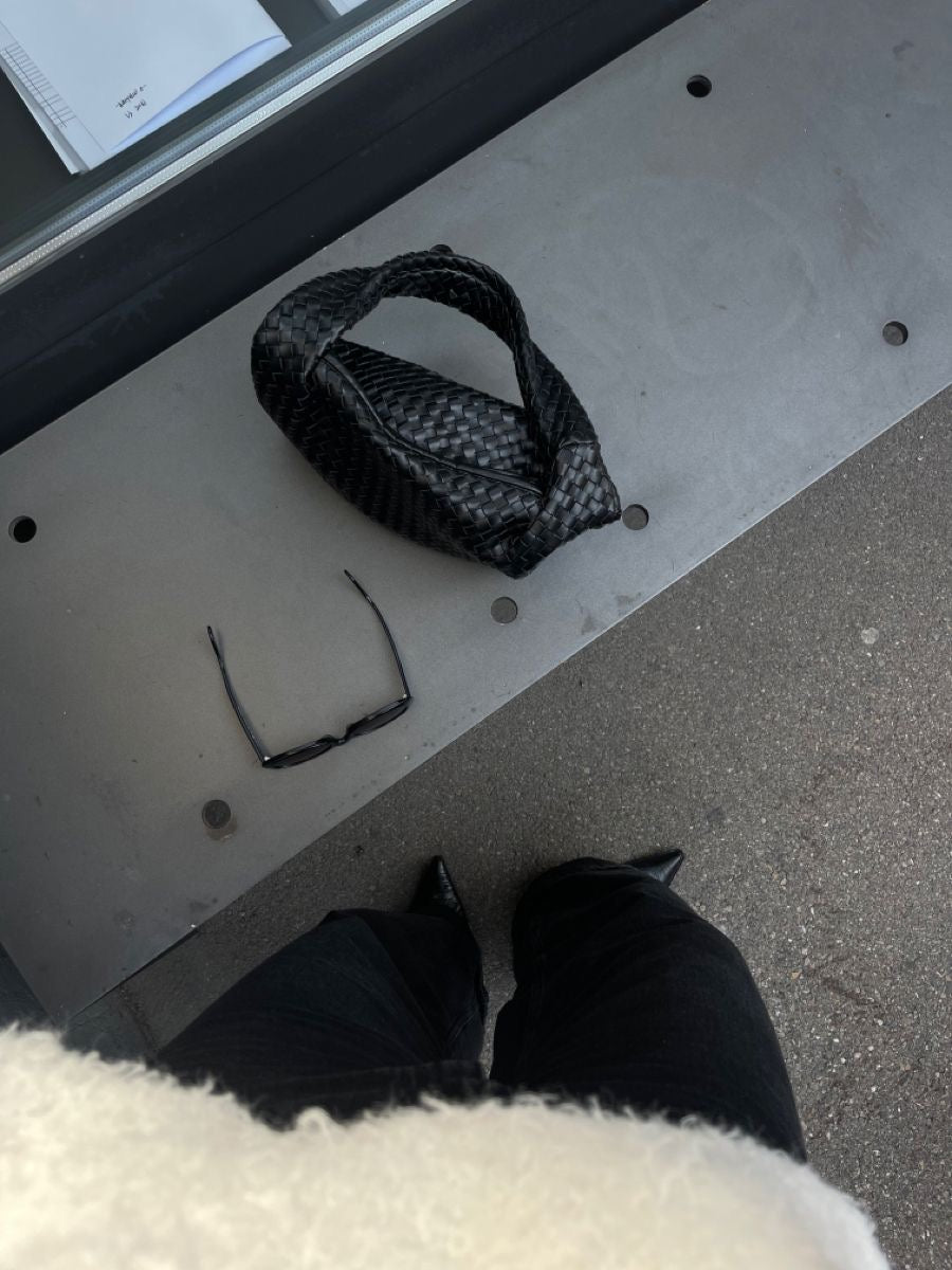 Person wearing black pants and a white coat standing on a metal surface with a woven bag in black colour.