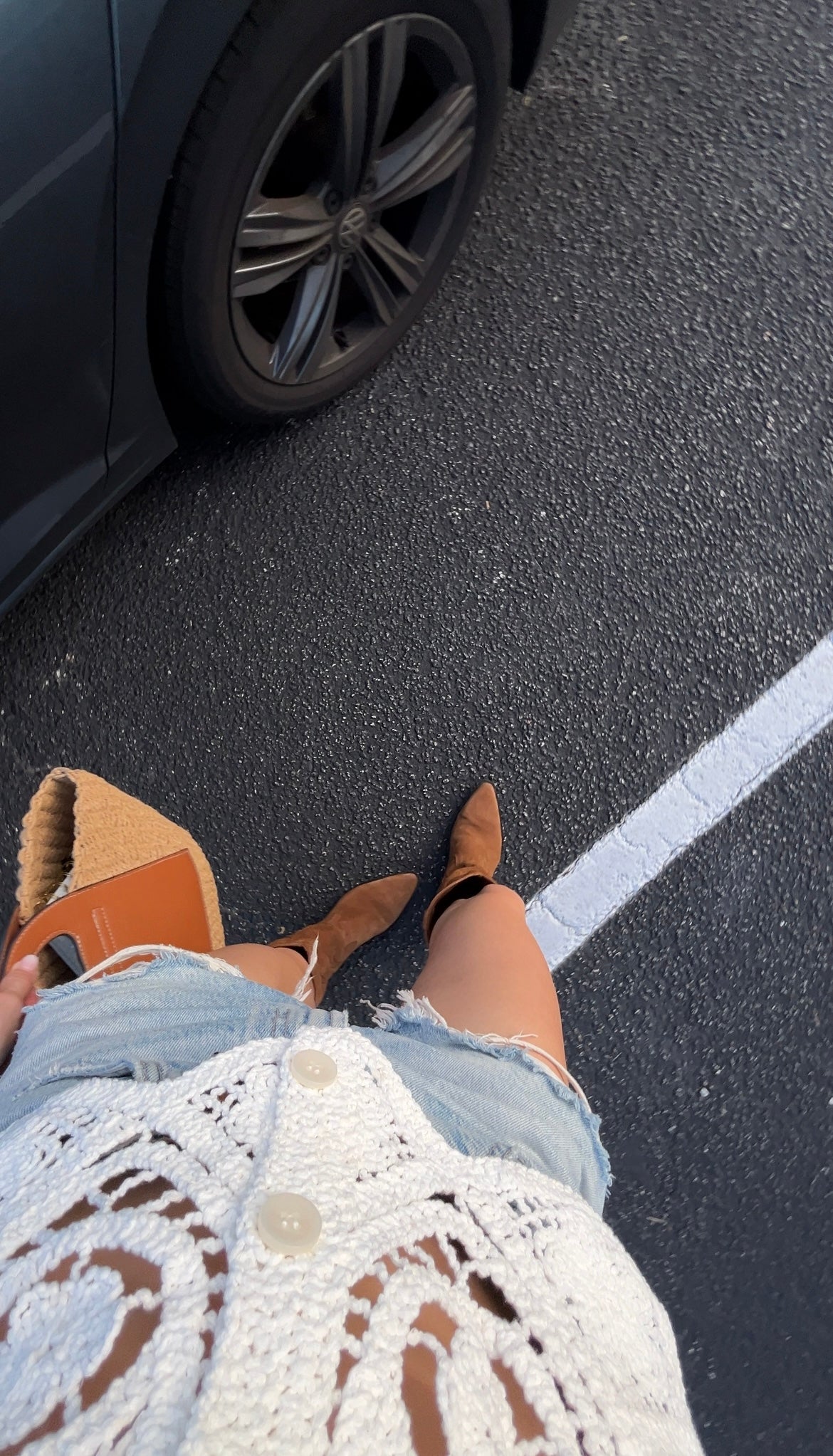 Person wearing brown shoes and light blue shorts standing on a road next to a car capturing a minimalist shot of a beach bag