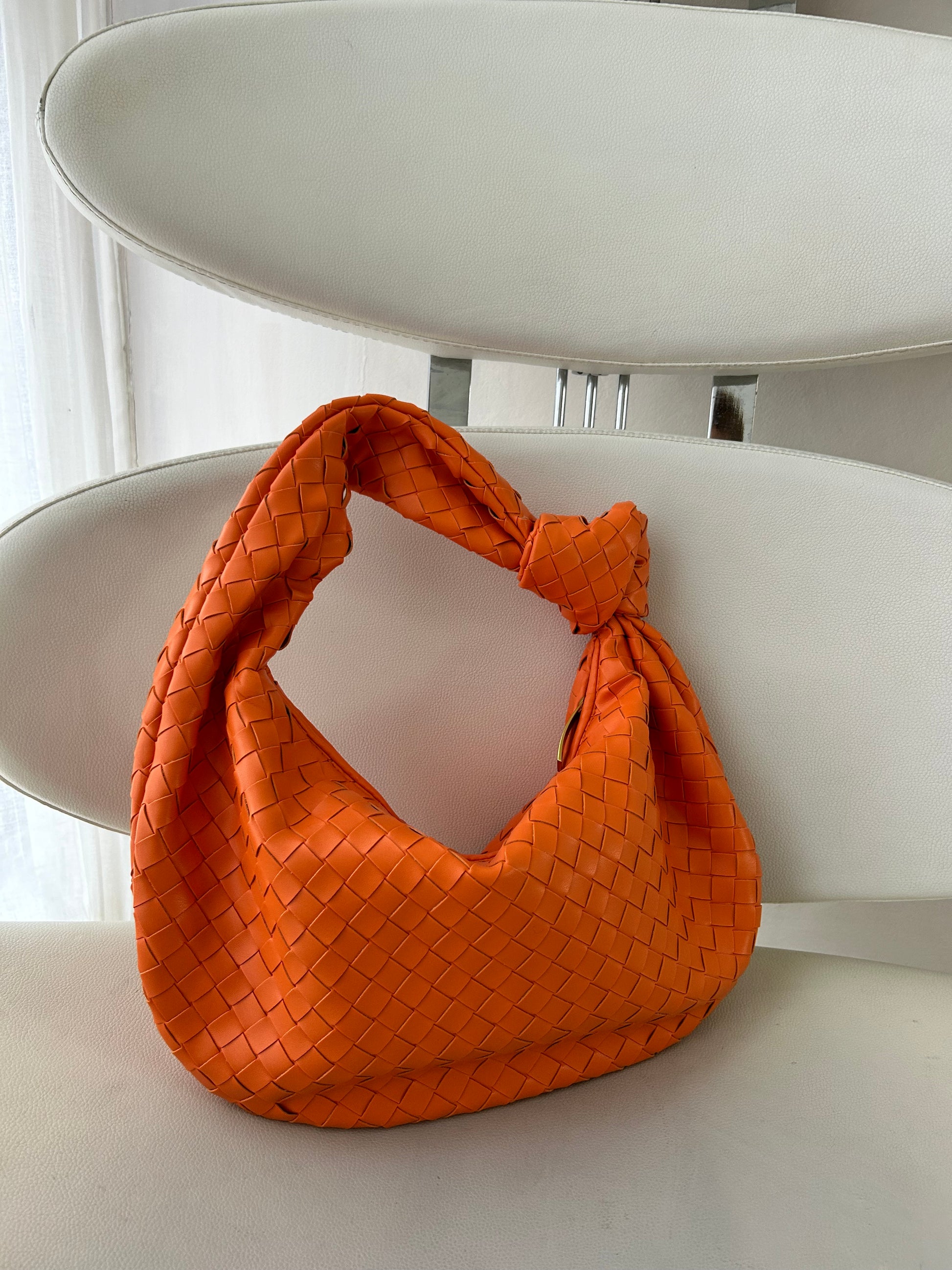 Large orange woven leather handbag with a soft knotted handle and distinctive triangle zip, resting on a staircase railing. A bold, statement accessory with sculptural texture and functional size.