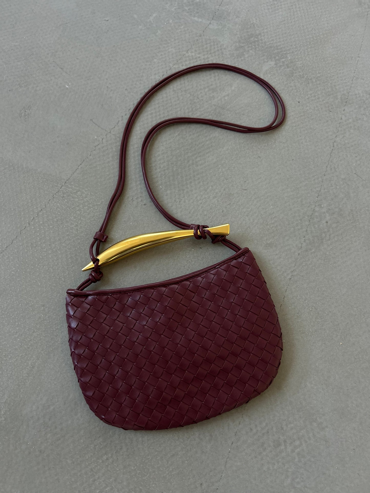 burgundy red woven handbag with gold bar handle detail in premium vegan leather placed on a concrete floor in grey colour