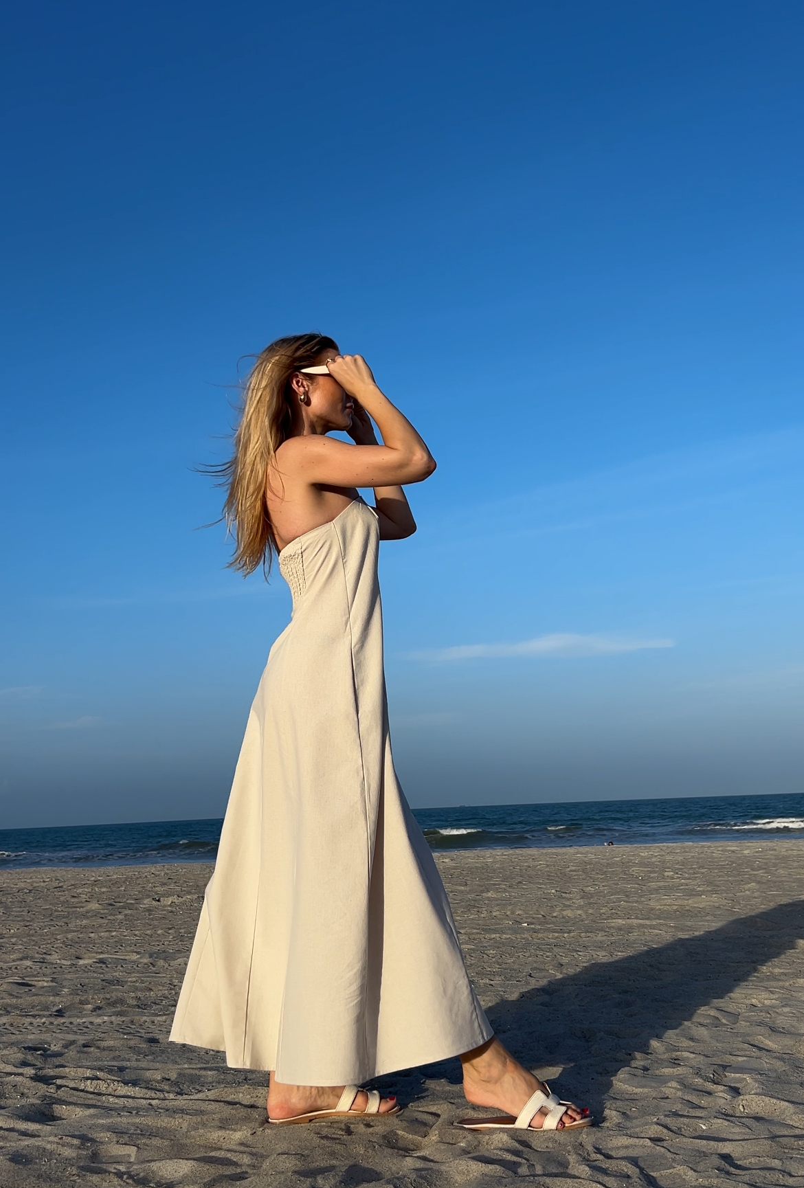 Woman in a long, light-colored linen dress standing on a beach with a clear blue sky.