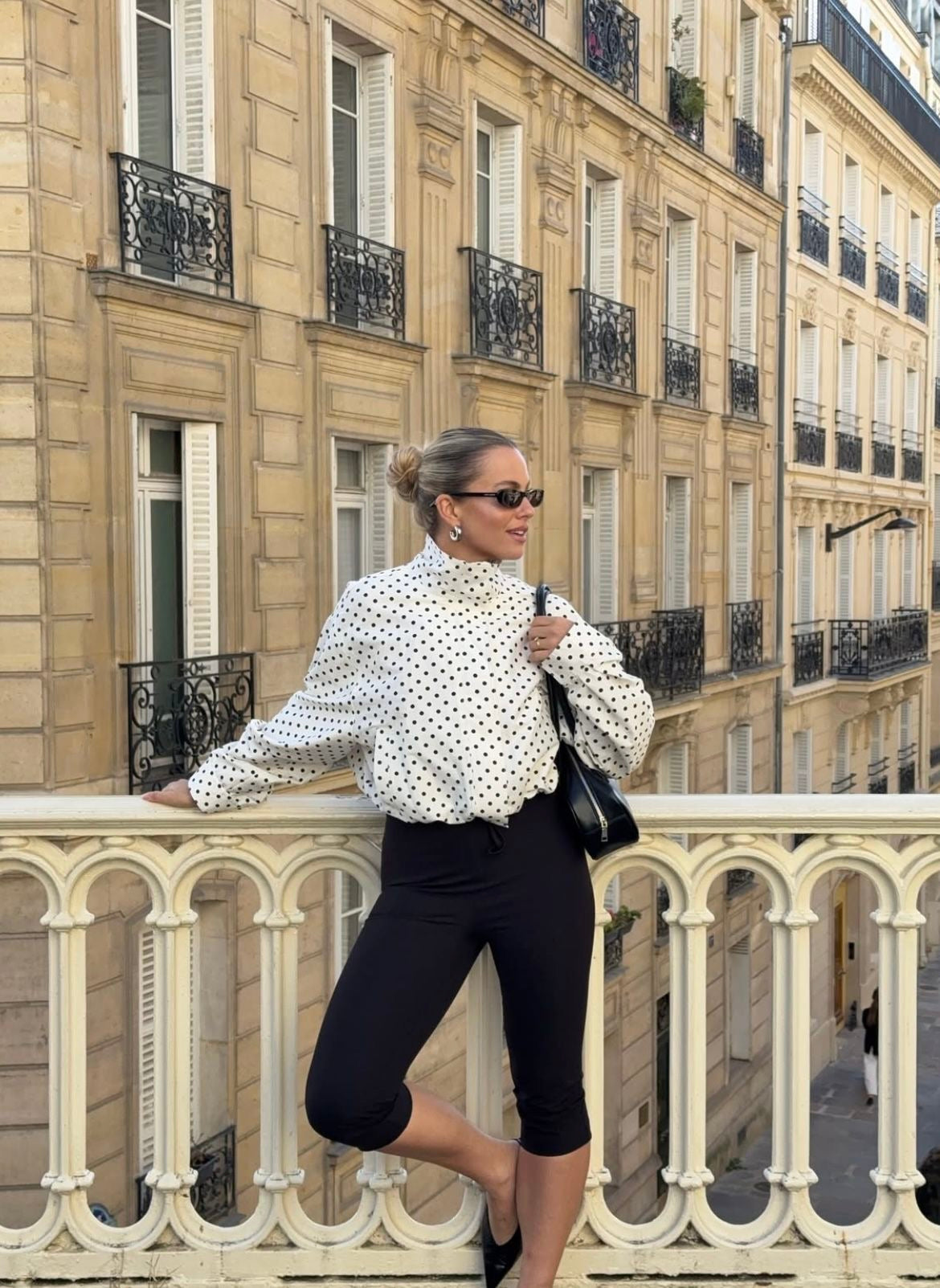 Woman in a polka dot viral bomber jacket and black pants standing on a balcony with classic building architecture in the background.