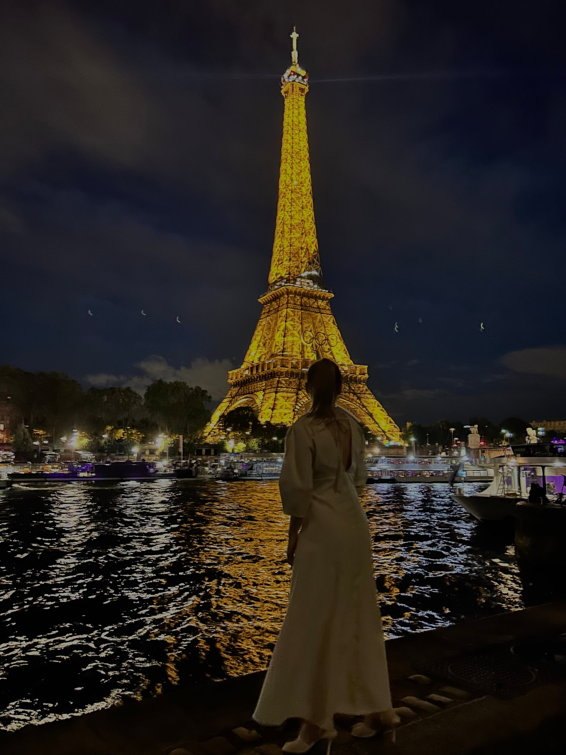 Woman in a white dress standing by a river with the Eiffel Tower illuminated in the background at night.