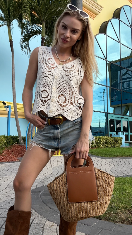 Woman in a white lace top and denim shorts holding a brown straw beach bag, standing in front of a colorful building in Florida