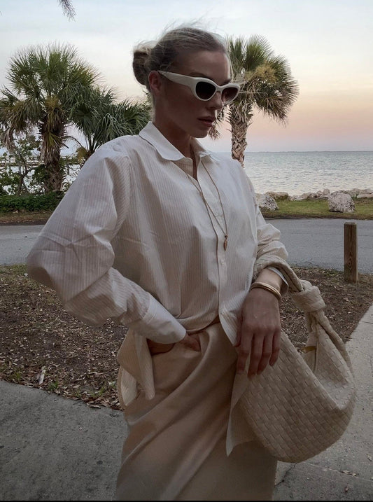 Woman in a white shirt and beige skirt standing by a beach with palm trees and ocean in the background, wearing cream woven handbag from PattyB