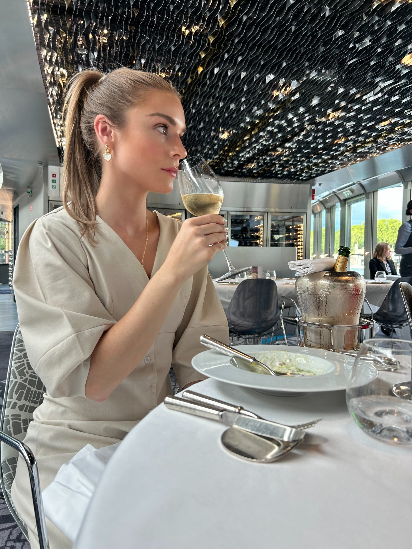 Woman sitting at a restaurant table, holding a glass of champagne wearing minimalist linen dress
