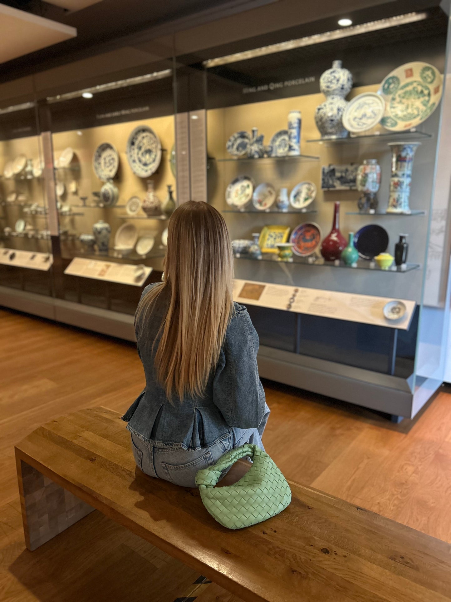 Woman sitting on a bench in a museum looking at ceramic plates displayed in cases.