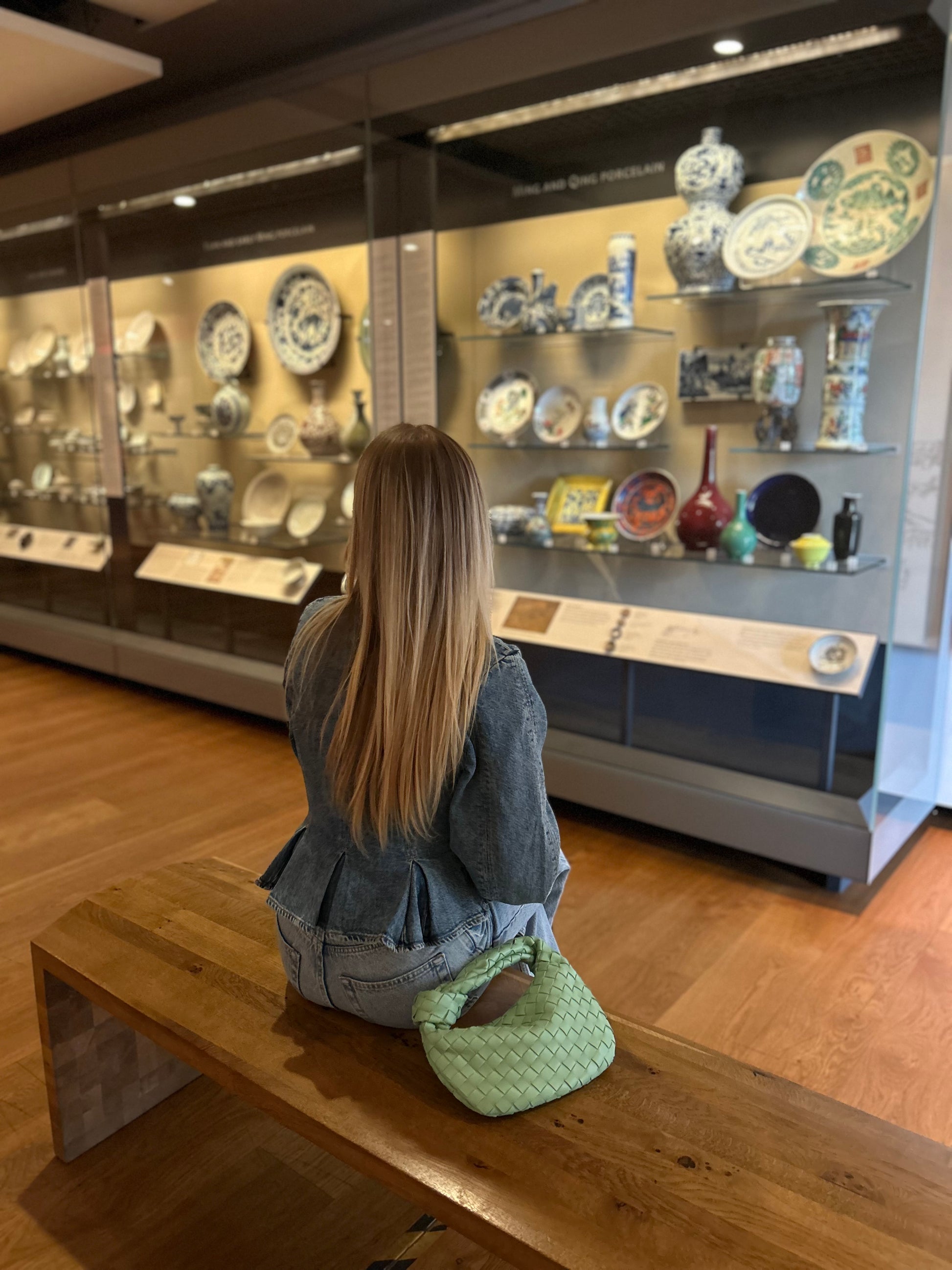Woman sitting on a bench in a museum looking at ceramic plates displayed in cases.
