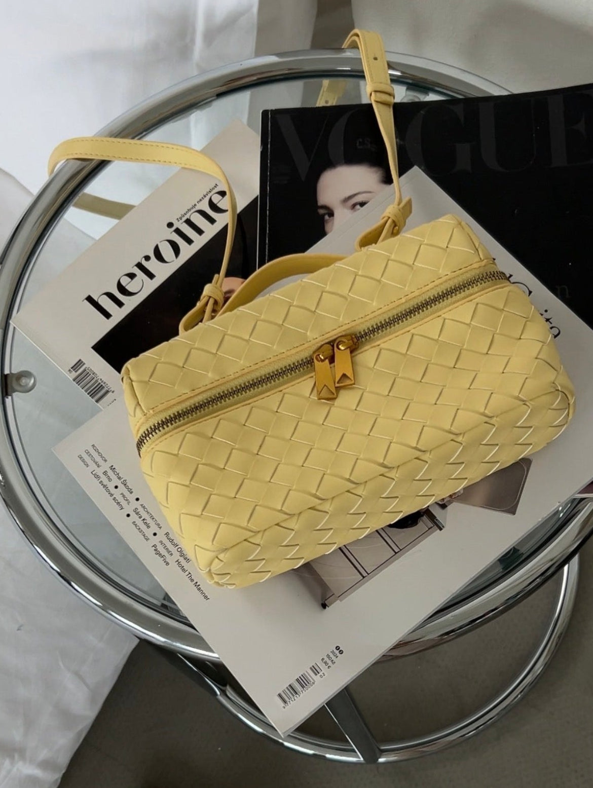 Yellow woven handbag on a glass table with a magazine underneath, next to a pair of slippers.