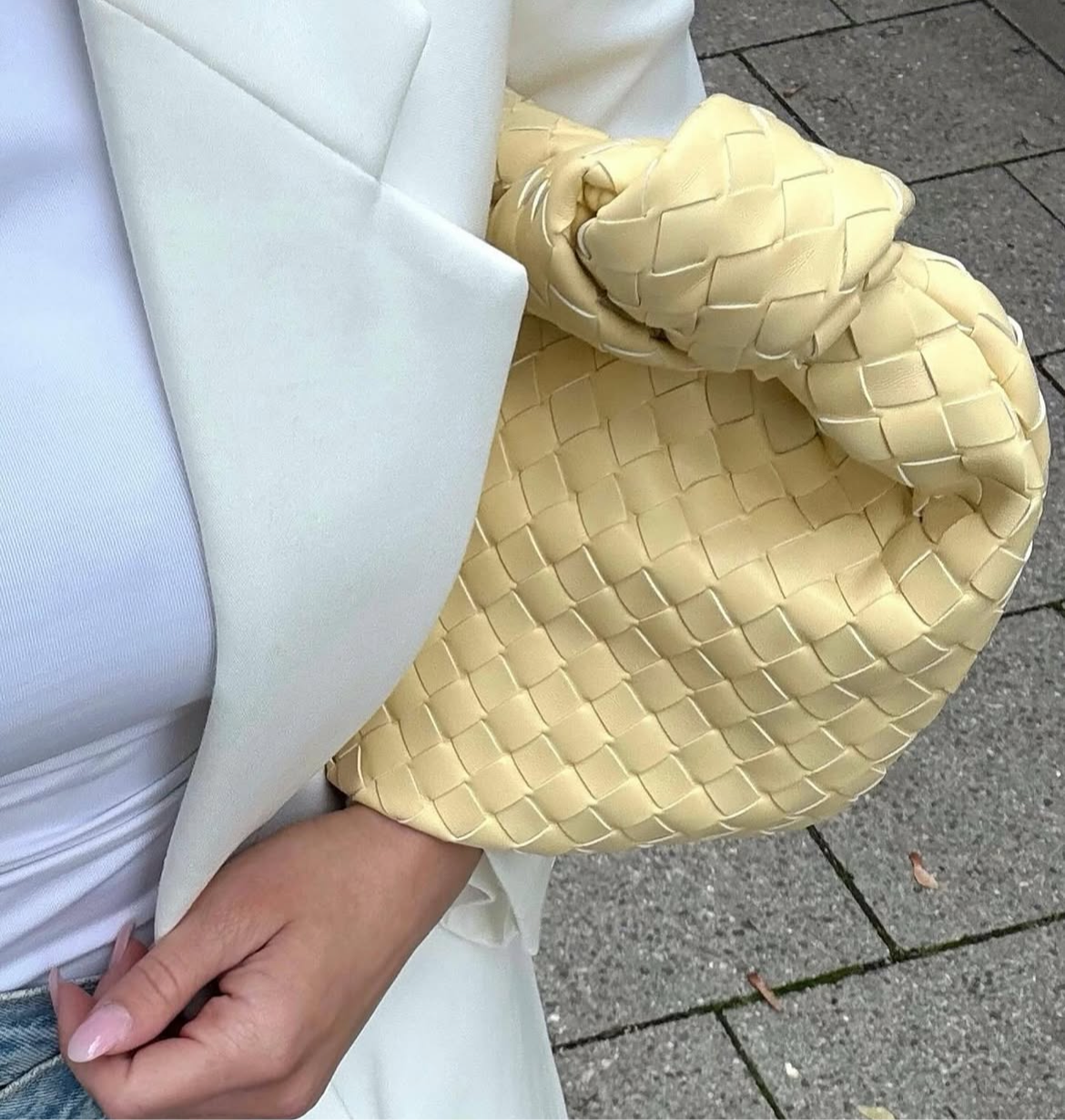 aesthetic shot of a pastel yellow woven handbag held by a person wearing a white outfit on a pavement background
