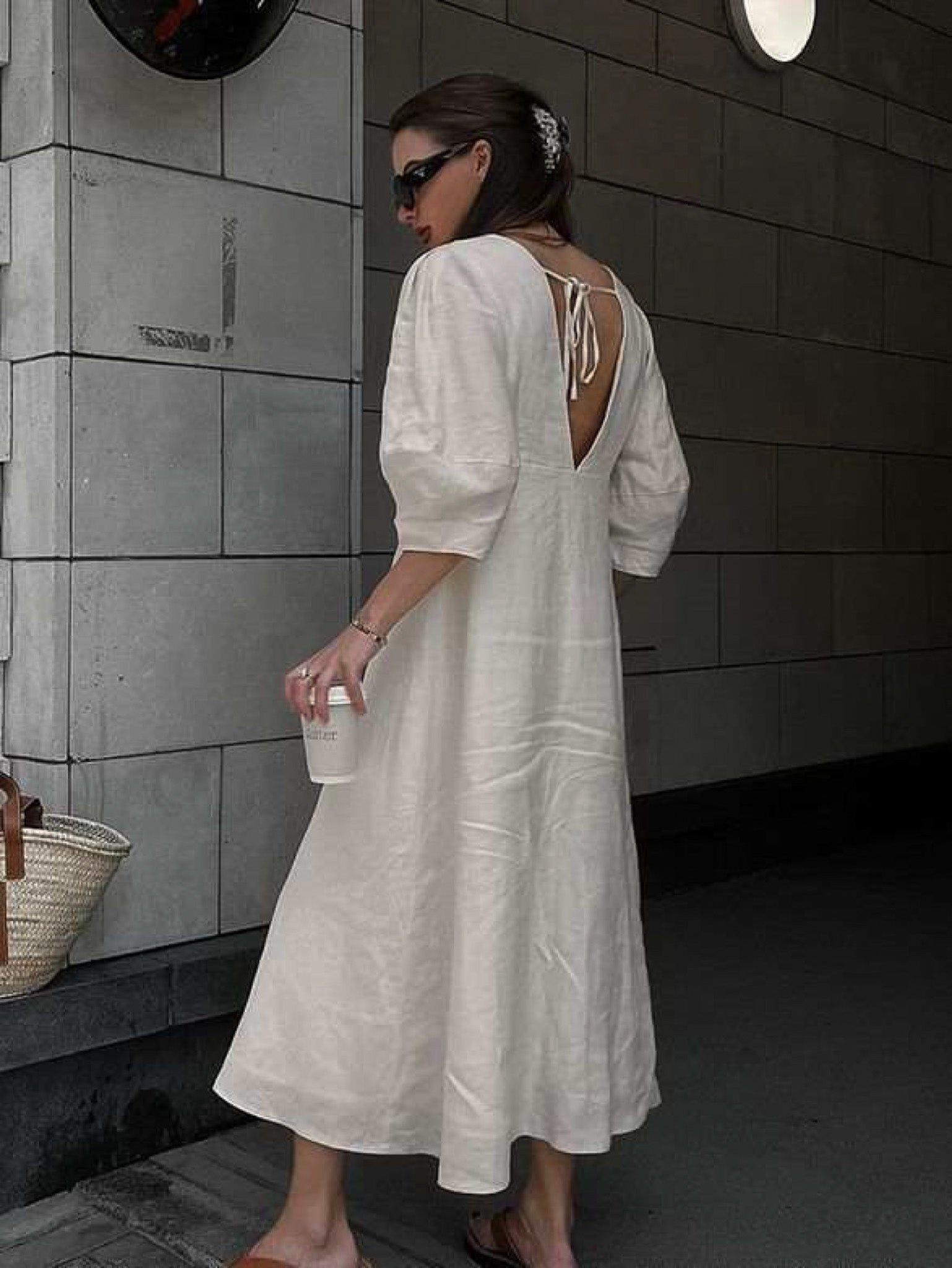 Back view of a woman wearing a cream linen midi dress featuring an open V-back with tie detail and elbow-length puff sleeves. The dress has a relaxed A-line silhouette and is styled with brown slide sandals and black sunglasses, creating a chic, effortless summer look.