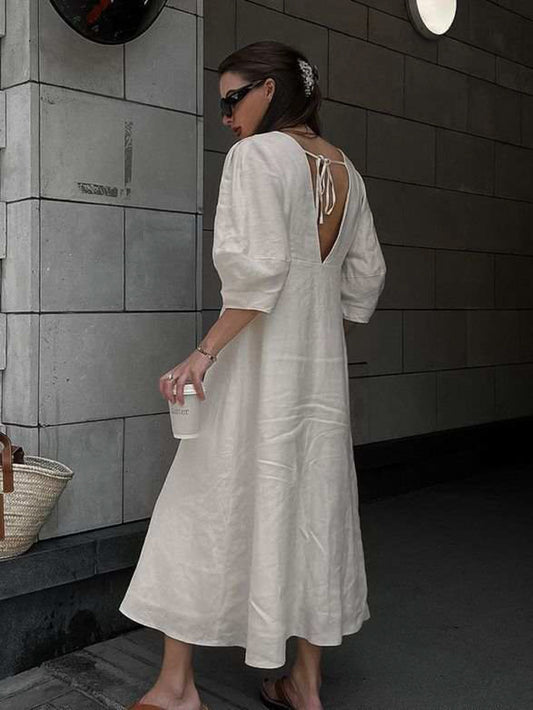 Back view of a woman wearing a cream linen midi dress featuring an open V-back with tie detail and elbow-length puff sleeves. The dress has a relaxed A-line silhouette and is styled with brown slide sandals and black sunglasses, creating a chic, effortless summer look.