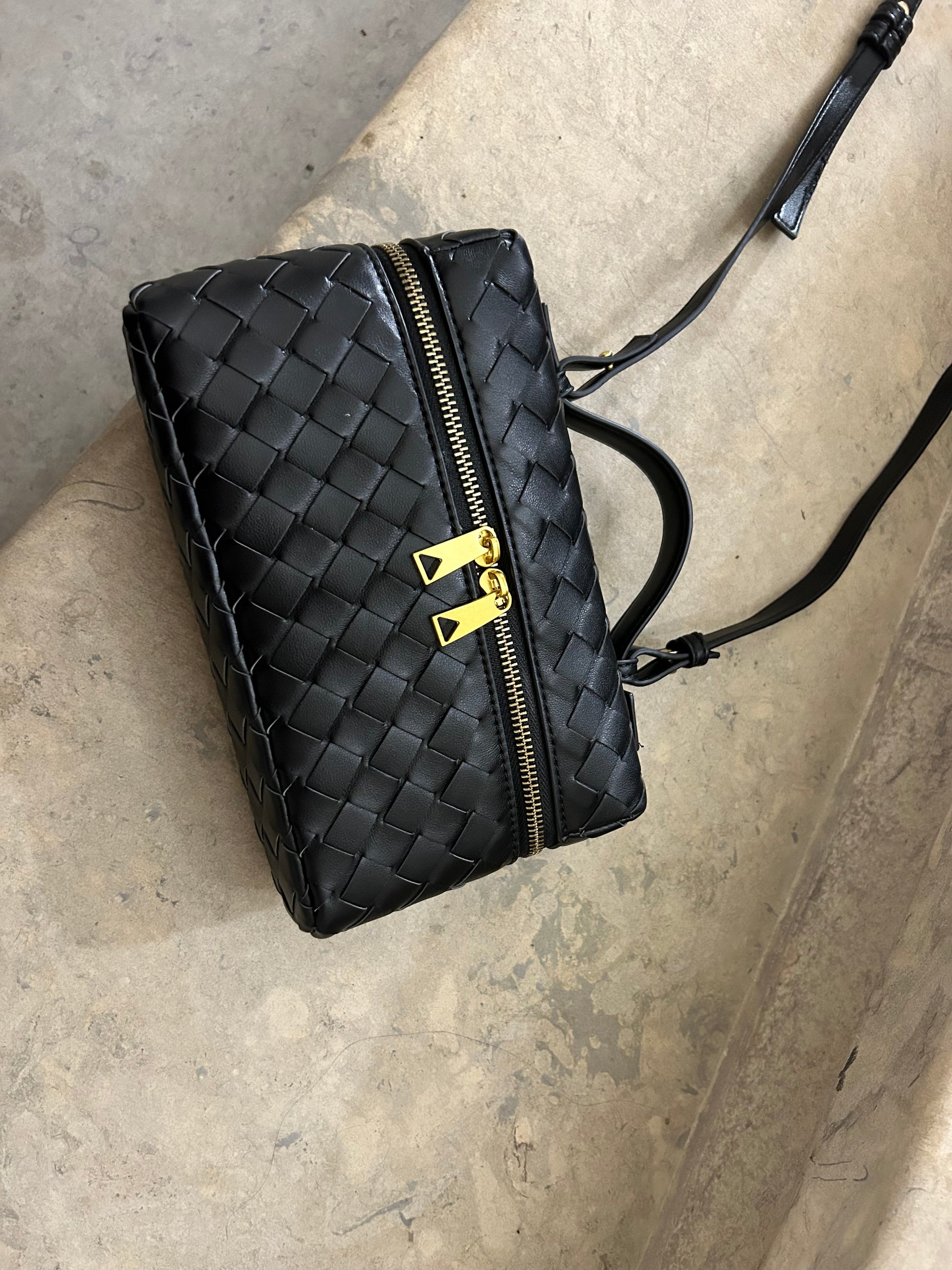 Black braided crossbody clutch elegantly placed on stairs, showcasing its intricate woven texture and sleek design. This versatile bag is perfect for adding a touch of sophistication to any outfit, making it a stylish accessory for any occasion.