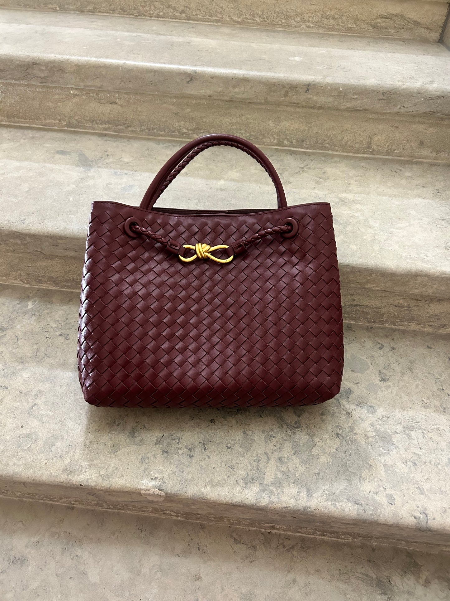A burgundy woven bag with a textured braided design rests elegantly on a set of stone stairs.The rich, deep red tone of the bag contrasts beautifully with the neutral stair background,showcasing its stylish and sophisticated appearance. designer dupe